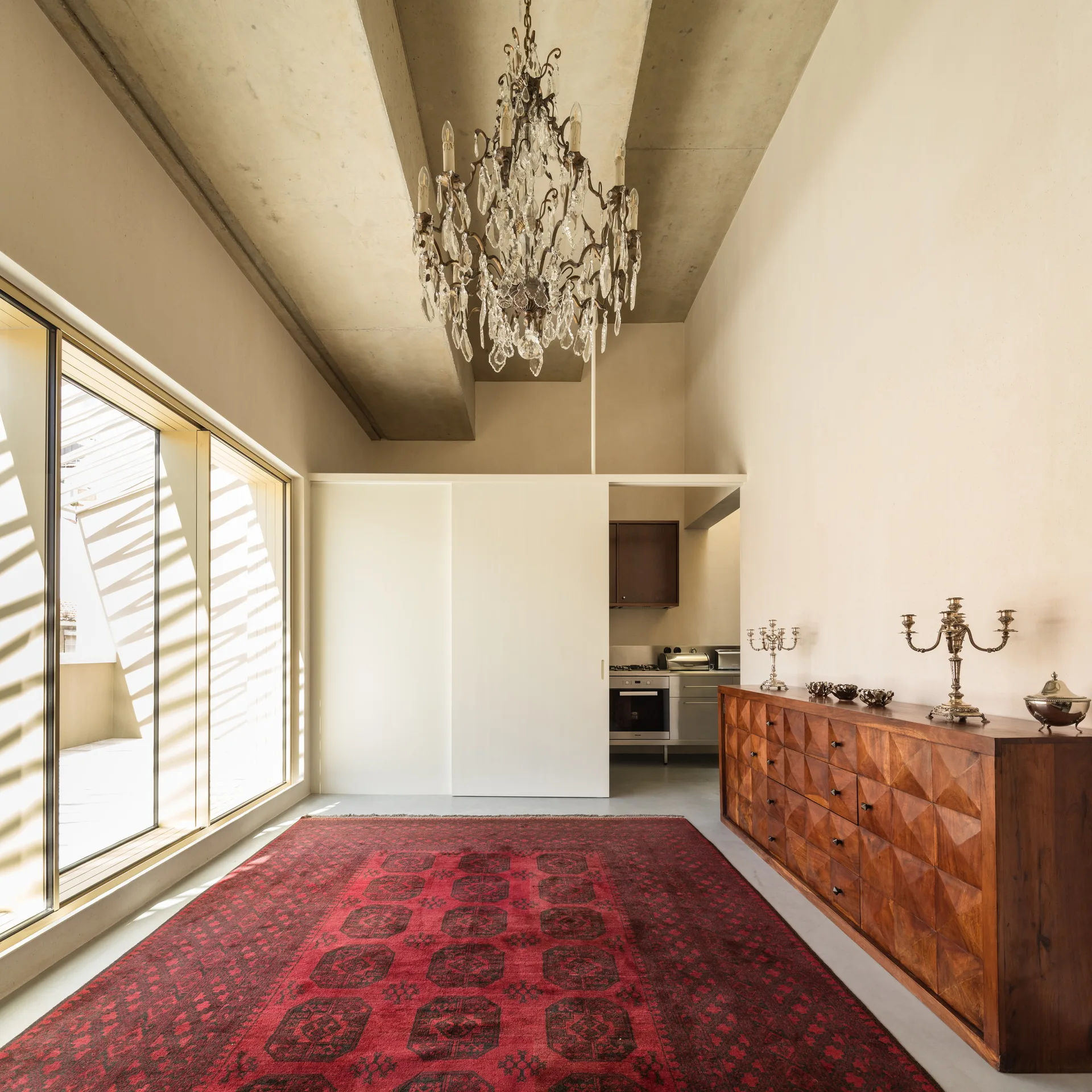A minimalist room features a large red patterned rug, a geometric wooden credenza, and an exposed concrete ceiling with a crystal chandelier.