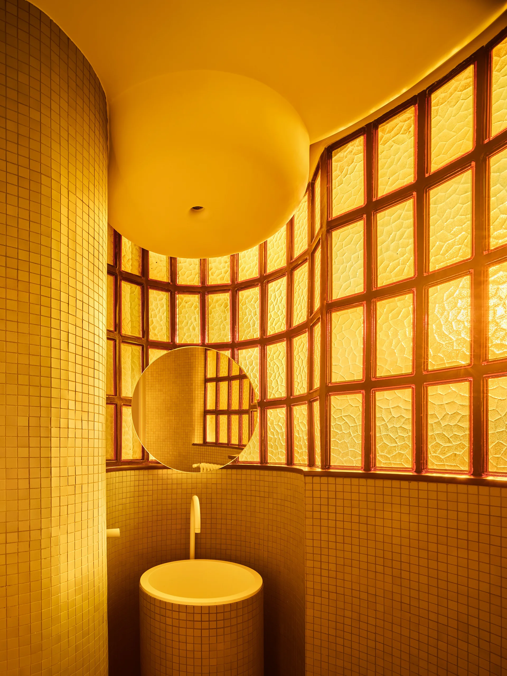 A minimalist yellow powder room features a curved glass block wall, a circular mirror, and a cylindrical tiled pedestal sink.