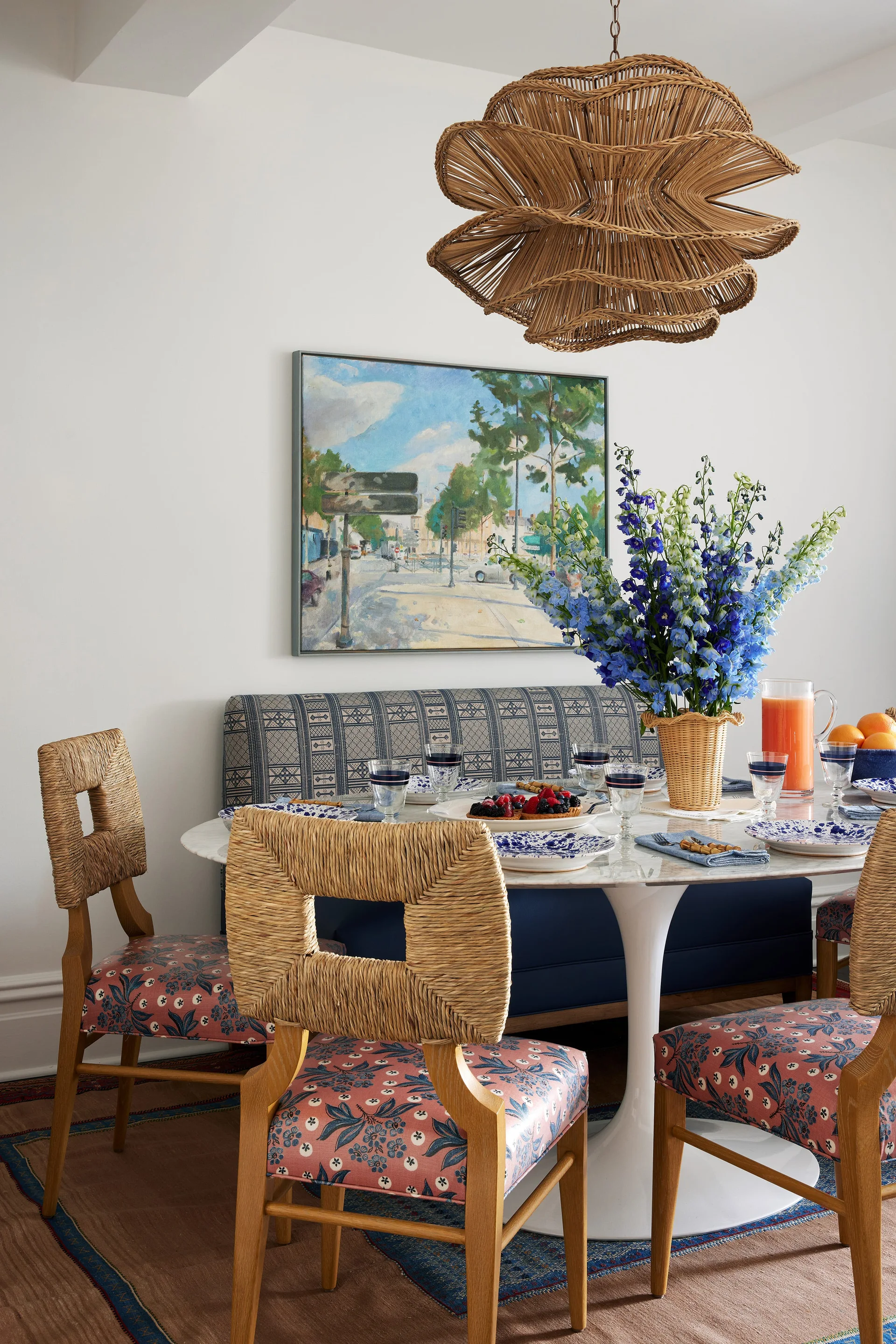 A dining room featuring a round pedestal table, floral-print chairs, and a blue patterned banquette under a sculptural flower-shaped wicker pendant light.