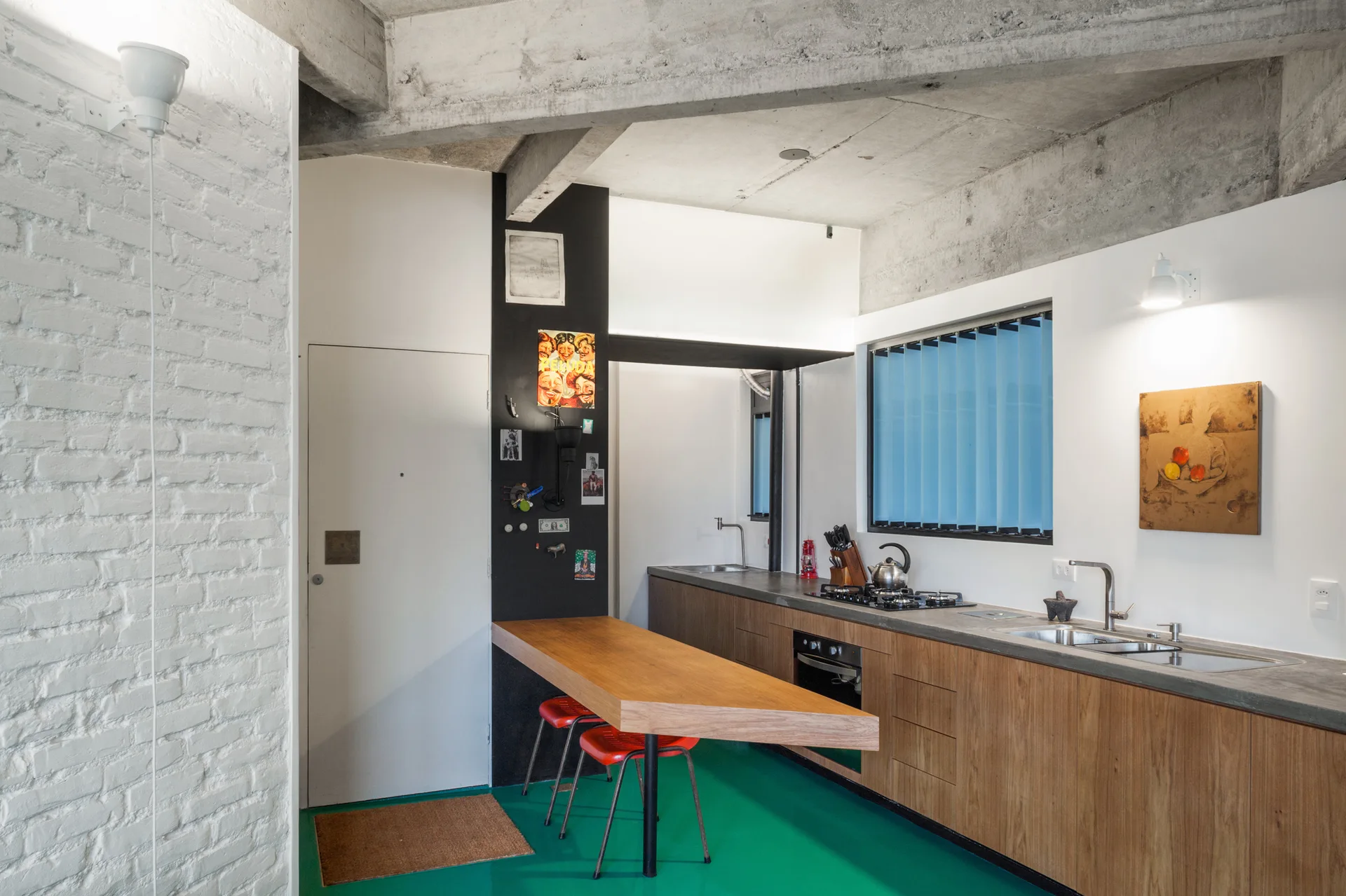 An industrial kitchen features wooden cabinetry, an angled wooden breakfast bar, red stools, a bright green floor, and an exposed concrete ceiling.