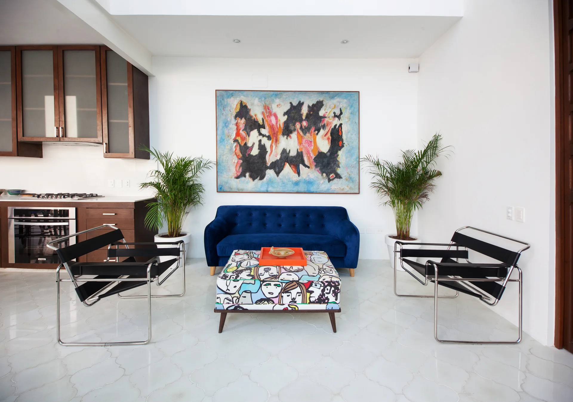 A blue tufted velvet sofa and two black leather armchairs surround a graphic illustrated coffee table on a white tile floor.