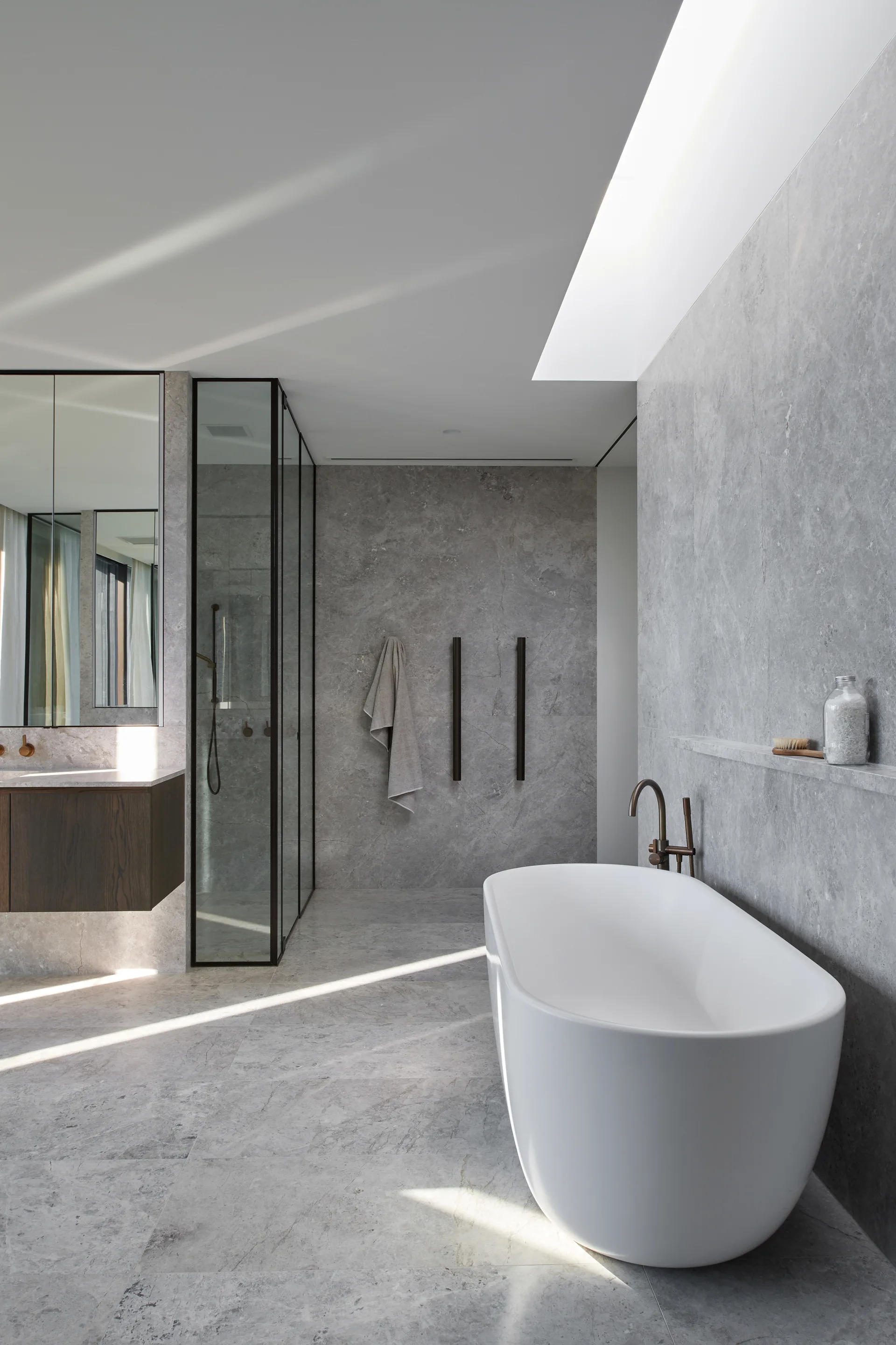 A minimalist bathroom featuring grey stone tiled walls and floors, a white freestanding bathtub, and a glass-enclosed walk-in shower.