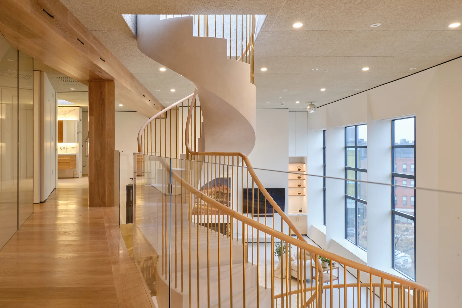 A modern loft interior with a central curved plaster spiral staircase featuring brass vertical spindles and a wood handrail next to a glass mezzanine railing.