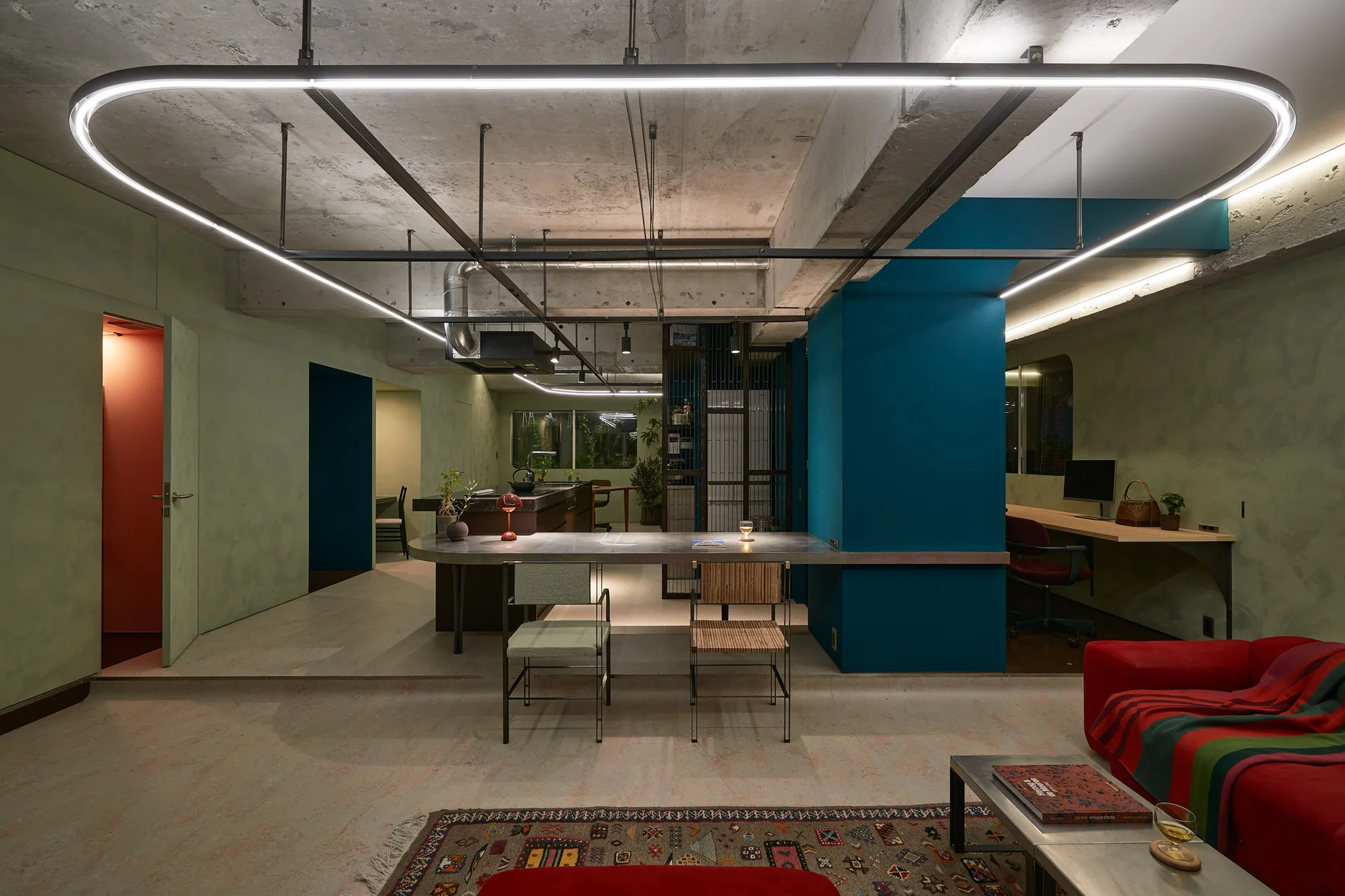 An industrial open-plan living area features sage green walls, an exposed concrete ceiling with a large oval pendant light, and a metallic kitchen counter.