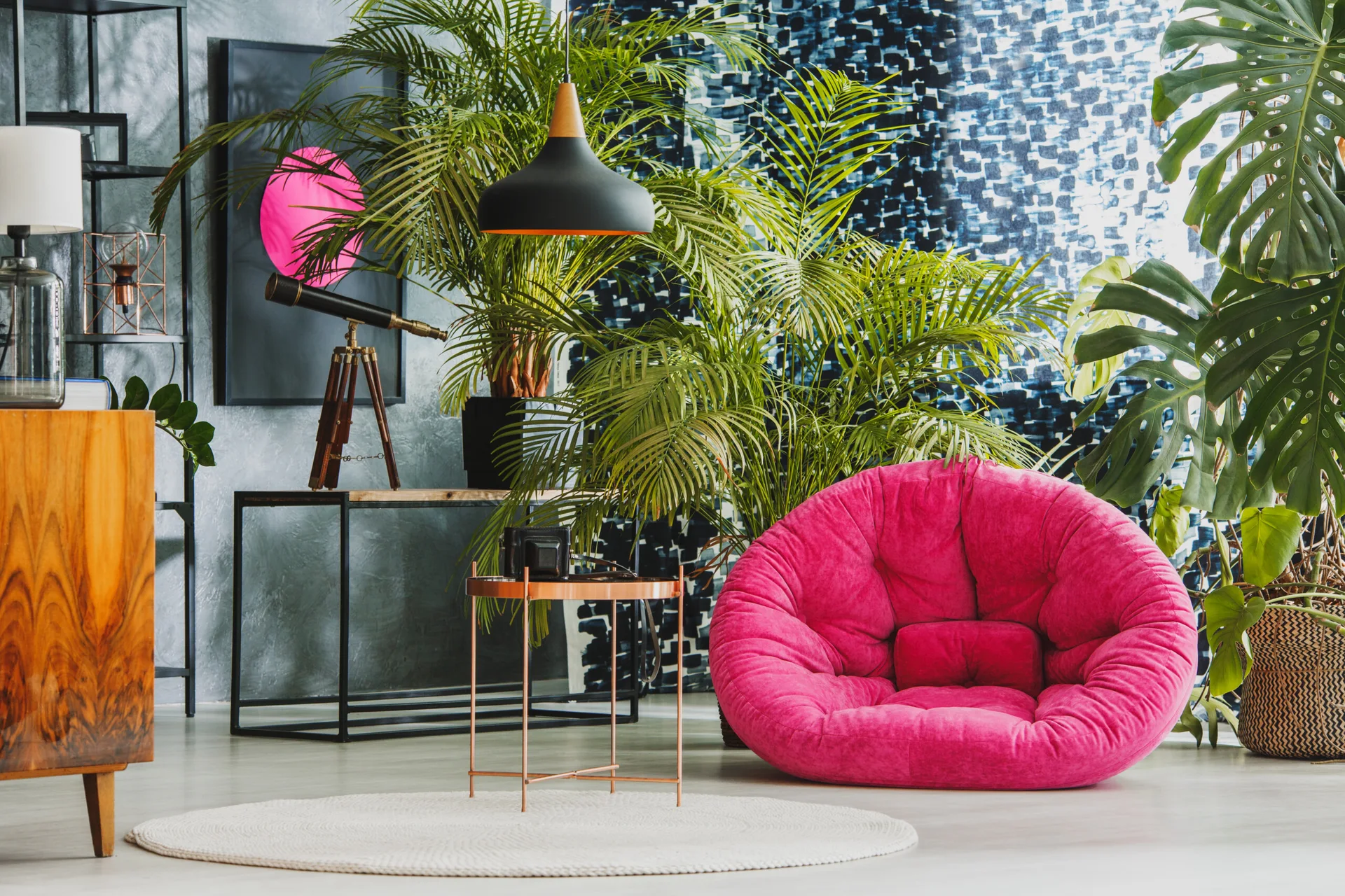 An eclectic living room corner featuring a bright pink round chair, large potted plants, a telescope on a console table, and patterned curtains.