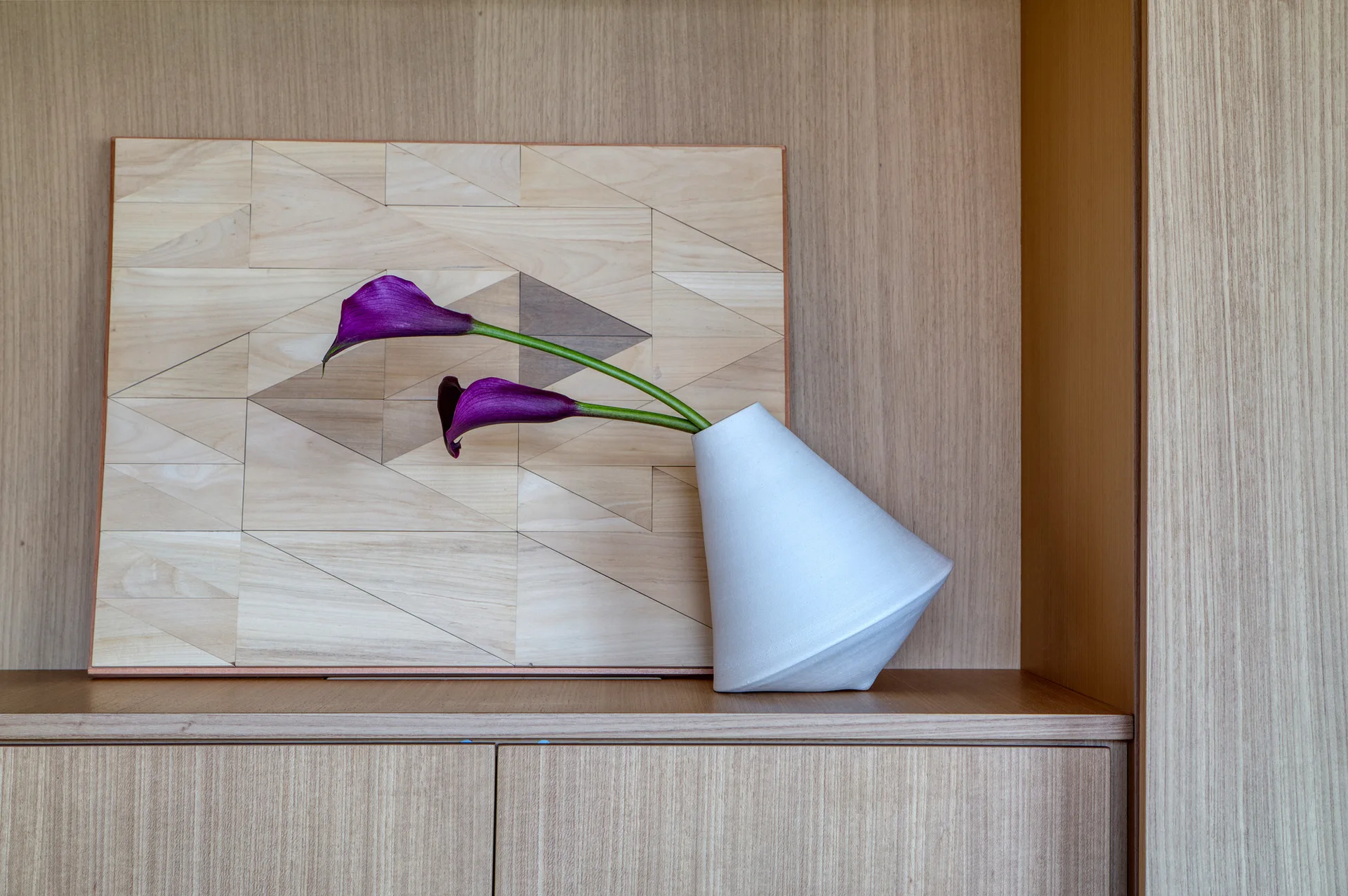 A white conical vase holding two purple calla lilies sits on a light wood shelf in front of geometric inlaid wood art.