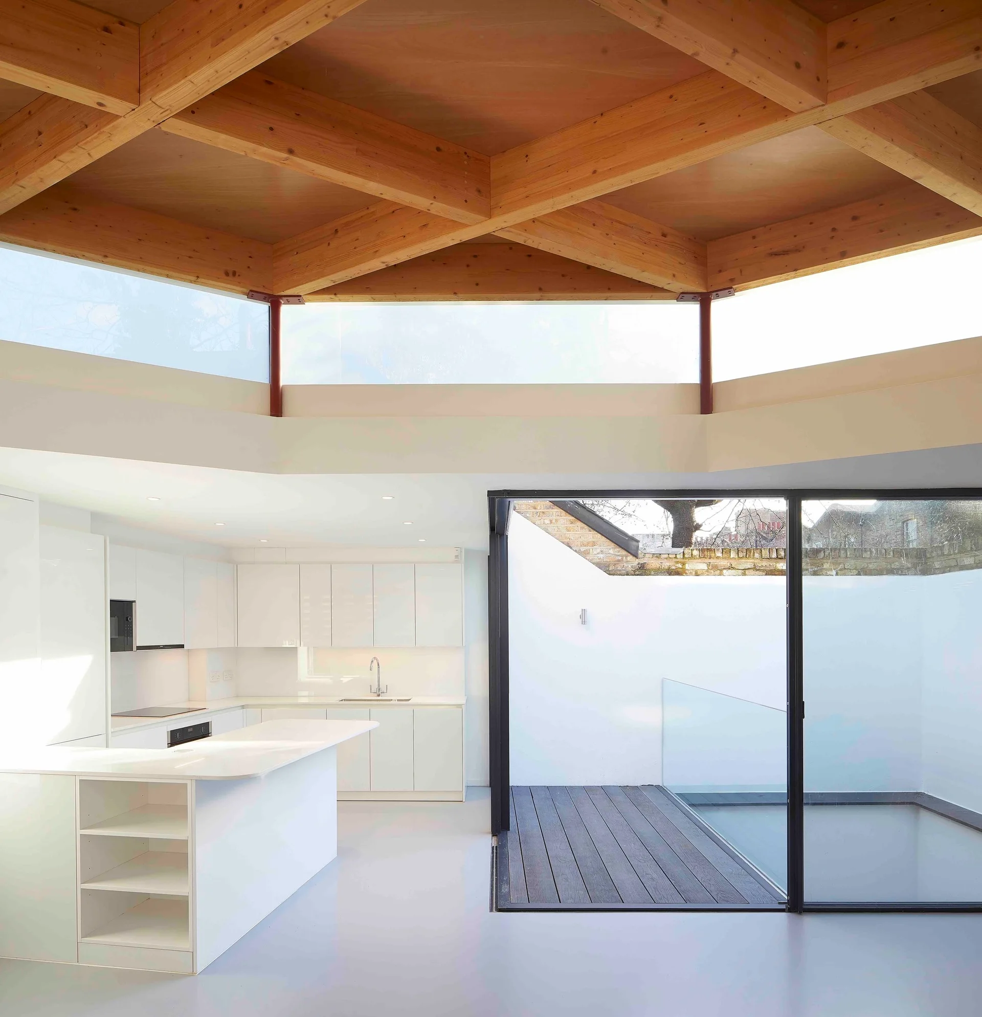 A modern white kitchen with minimalist cabinets, a central island, a geometric wood beam ceiling, and a large sliding glass door opening to a deck.