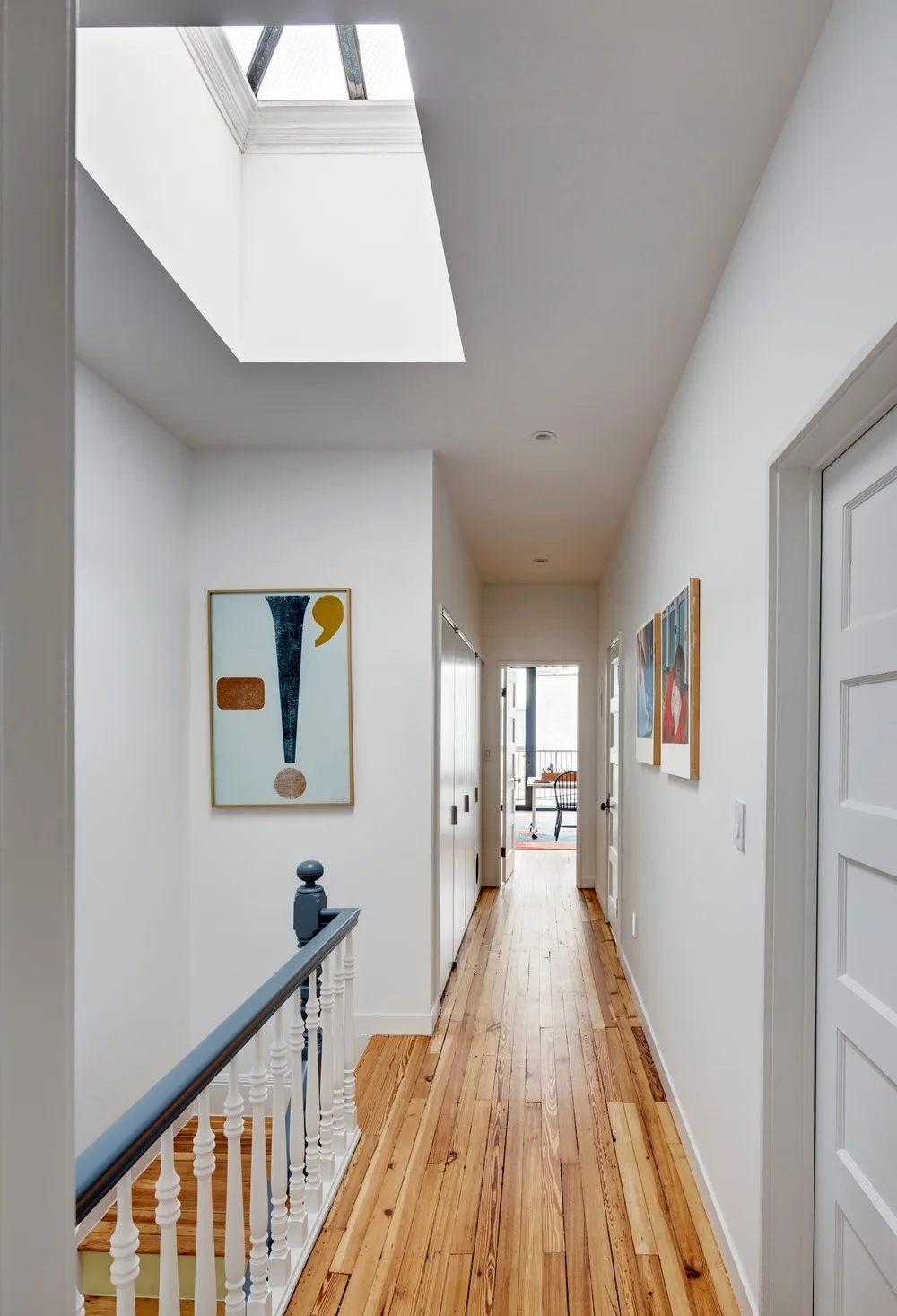 A white hallway featuring natural wood floors, a deep ceiling skylight, abstract art, and a staircase railing painted white with a blue handrail.