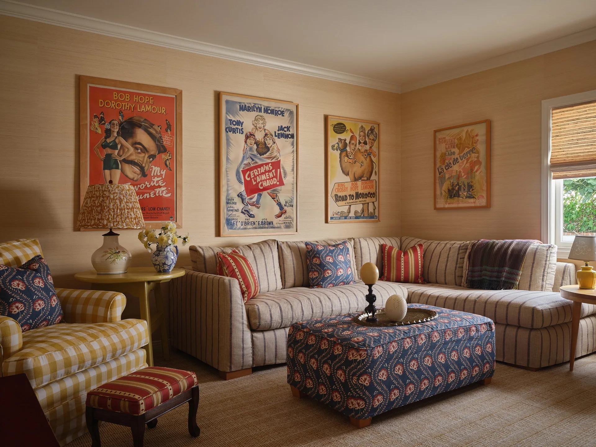 A living room features a striped sectional sofa, a patterned blue ottoman, and a checkered yellow armchair below framed vintage movie posters.