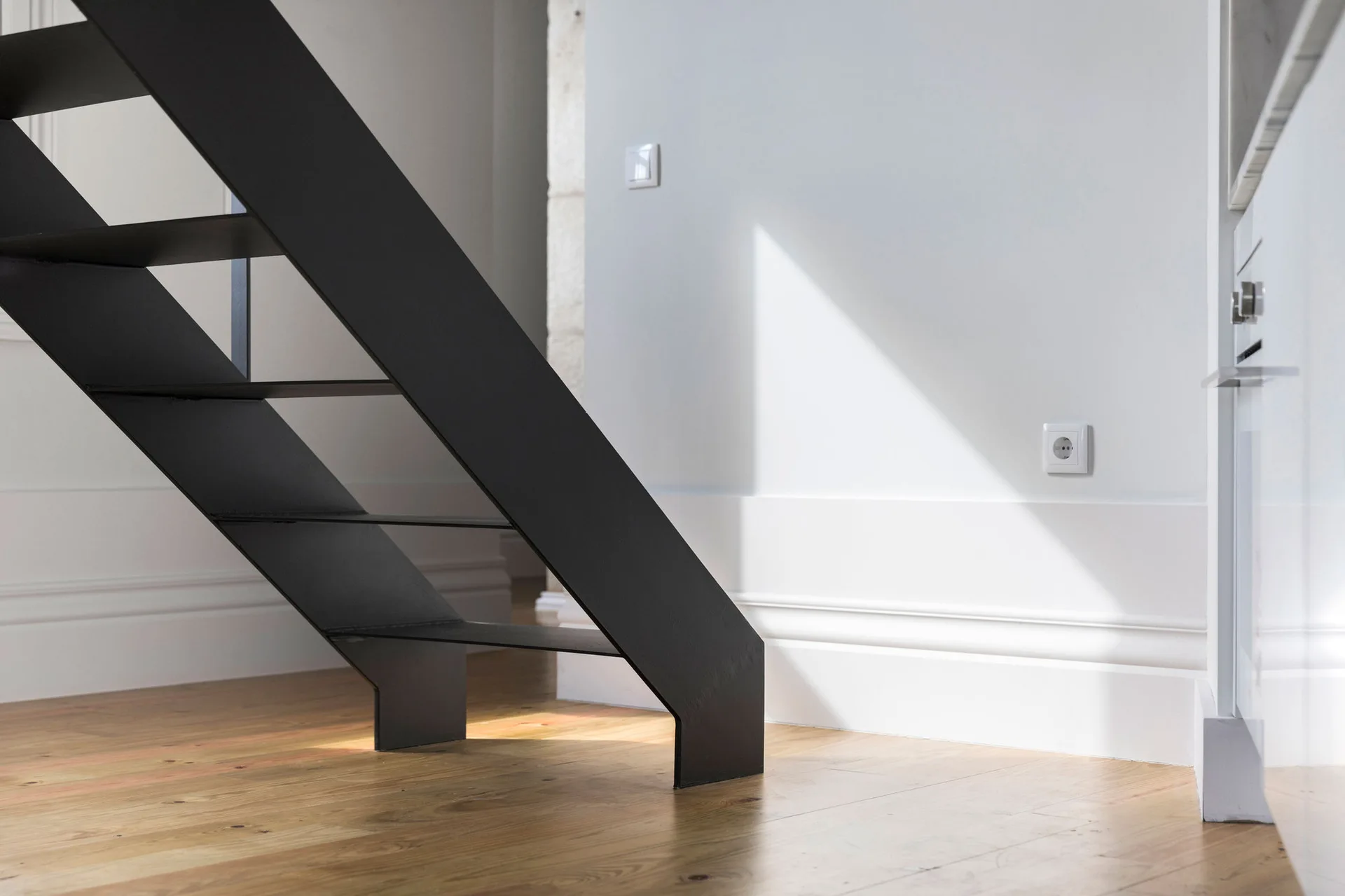 A modern black metal staircase rests on light wood plank flooring against a white wall with deep baseboards.