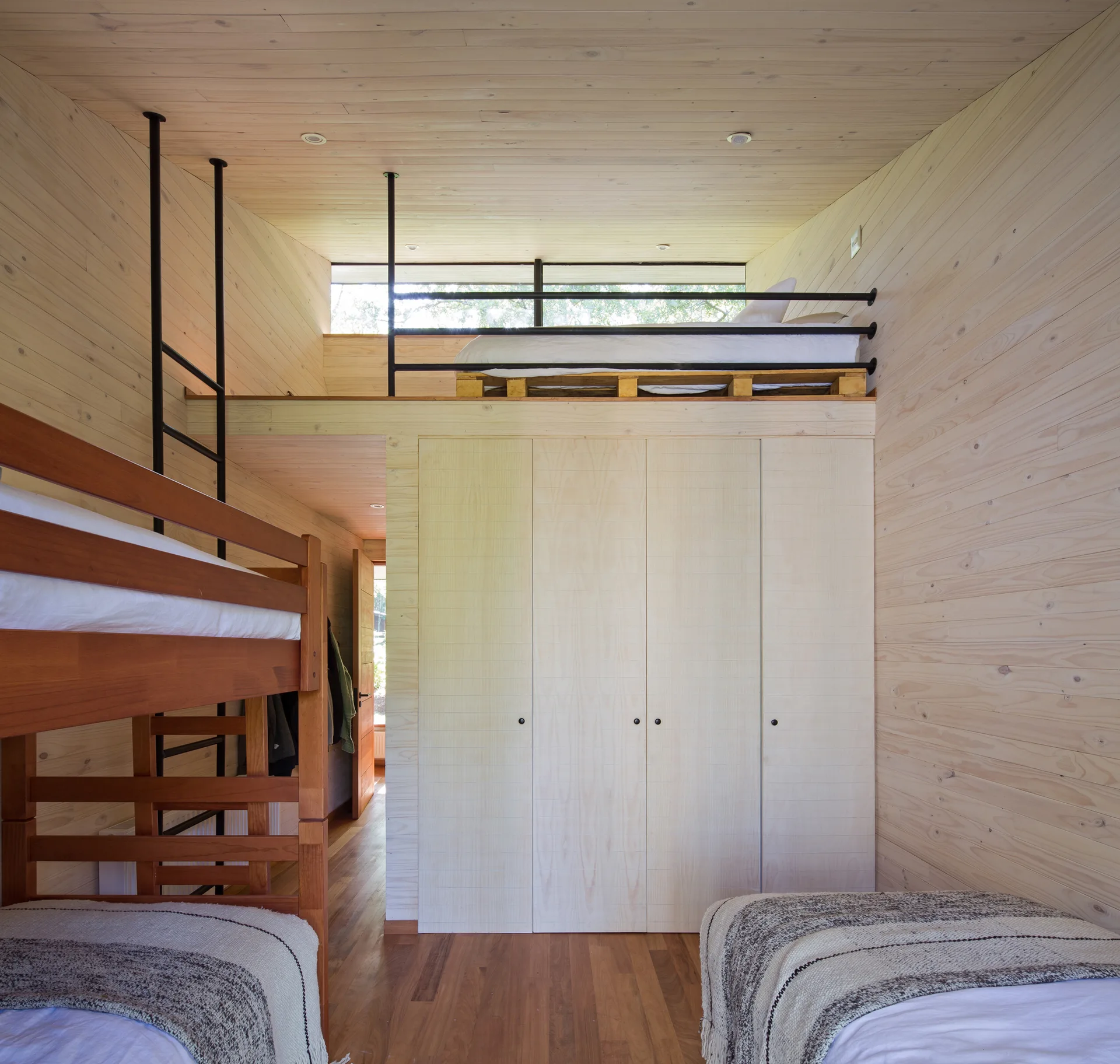 A light wood bedroom features a wooden bunk bed, a twin bed, and an elevated loft bed built above a light-colored wardrobe with a black metal railing.