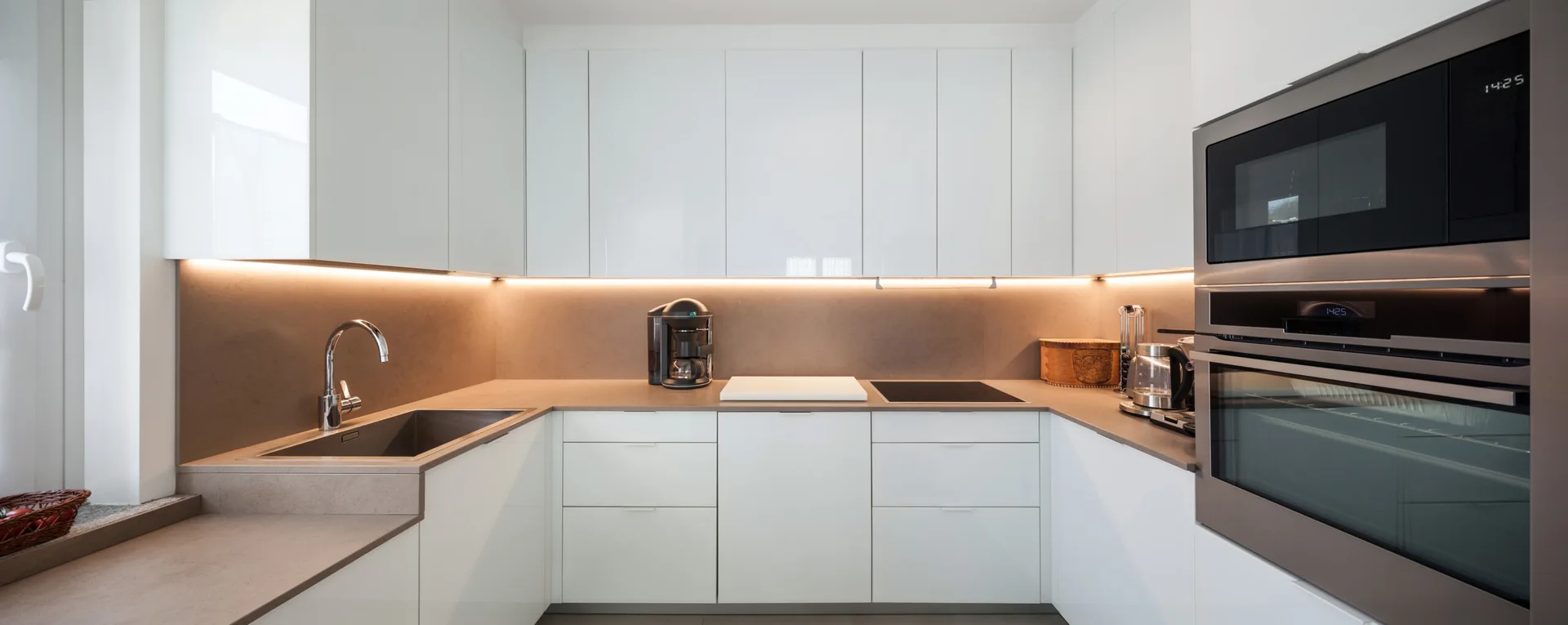 A modern U-shaped kitchen featuring glossy white handleless cabinets, matte grey stone-like countertops, and warm under-cabinet strip lighting.
