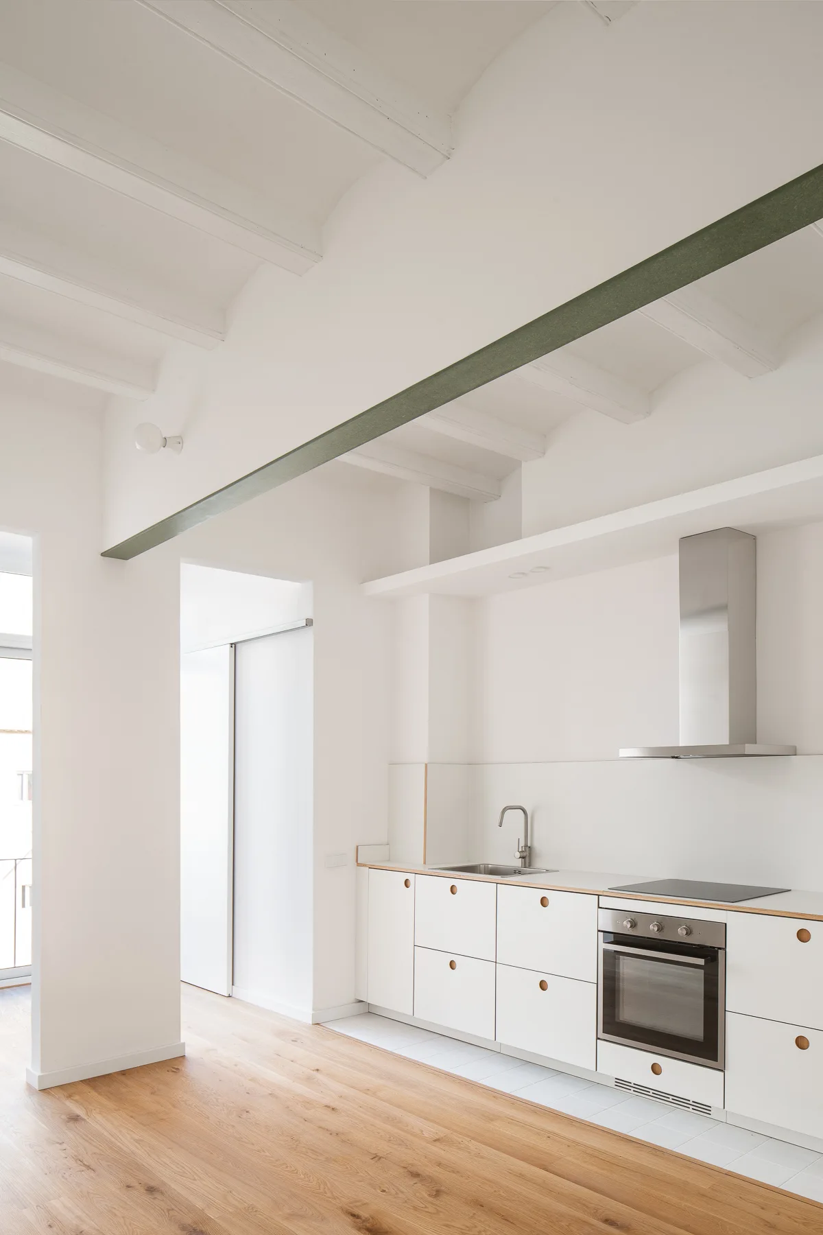 A minimalist white kitchen features flat-front base cabinets with circular cutouts, exposed white ceiling beams, and a dark green structural support beam.