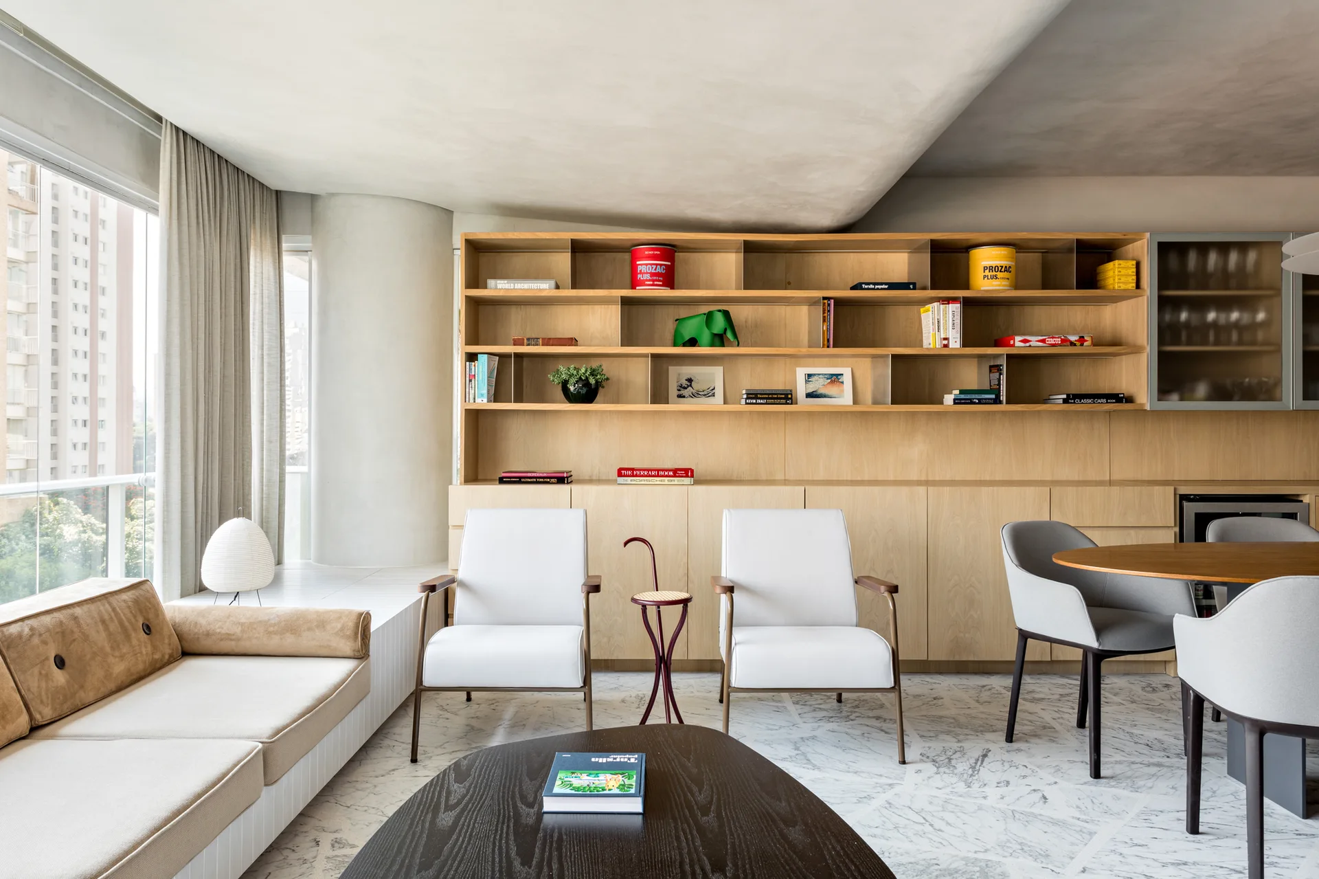 A modern apartment living room featuring a custom light wood wall bookshelf, white armchairs, and a veined marble floor.