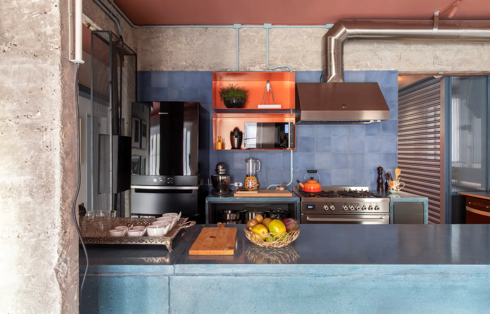 A modern residential kitchen features a blue tile backsplash, orange metal open shelves, a concrete island, and exposed industrial conduit.