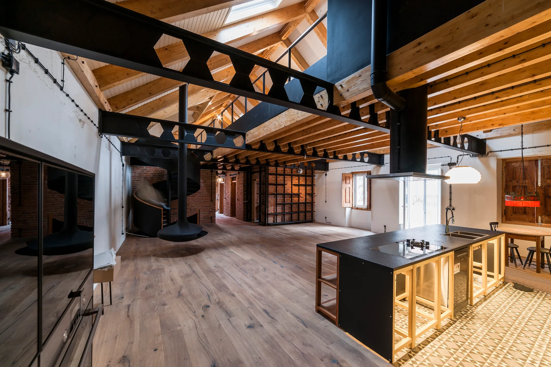 An open-plan loft kitchen features a black island on geometric tile, exposed wooden rafters, and a black spiral staircase.