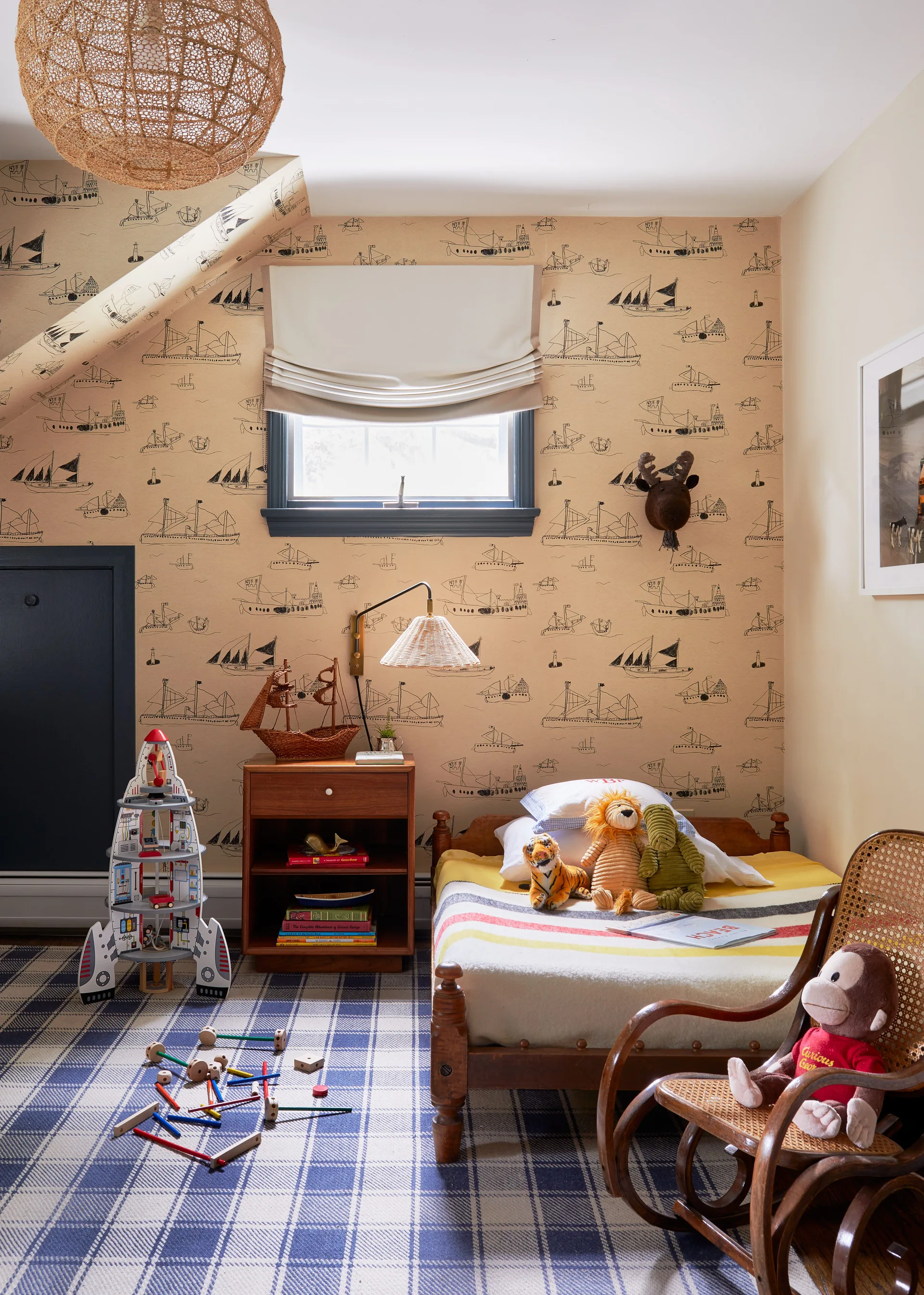 A kids bedroom featuring tan ship-patterned wallpaper, a wooden twin bed with stuffed animals, a blue plaid rug, and a small rocking chair.