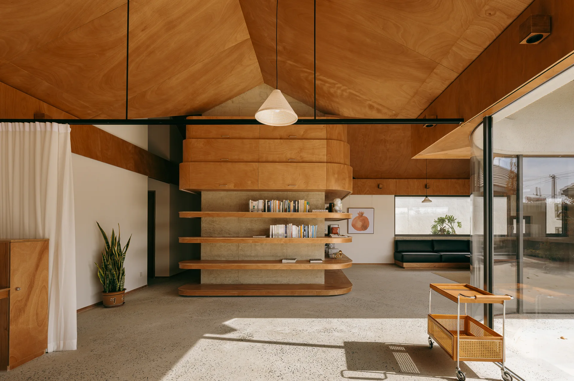 A minimalist living room features an angular plywood ceiling, a central curved wooden shelving unit, concrete floors, and a rolling cane bar cart.