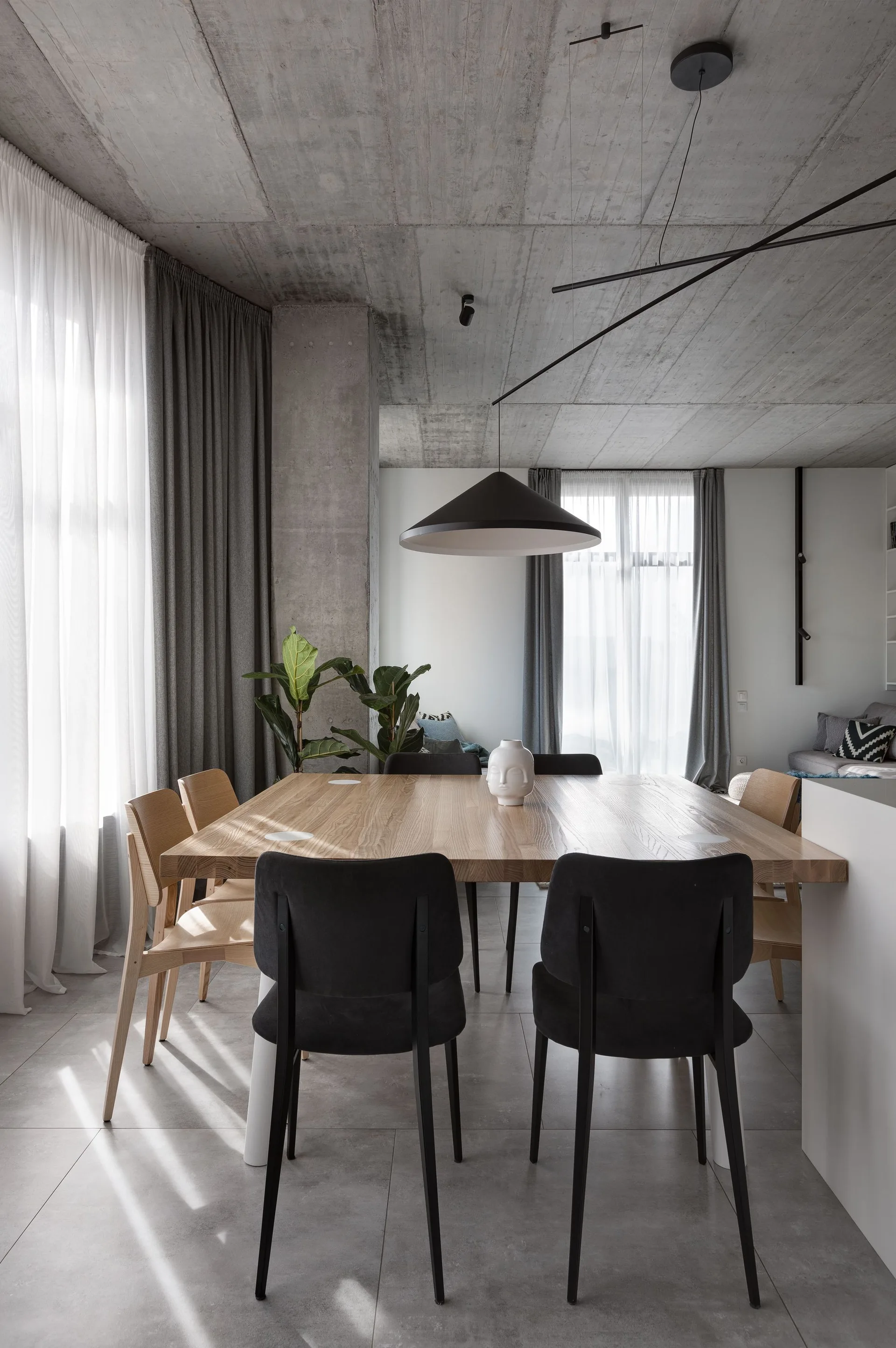 A modern dining room features a light wood table, black and wooden chairs, an exposed concrete ceiling, and a large black pendant light.