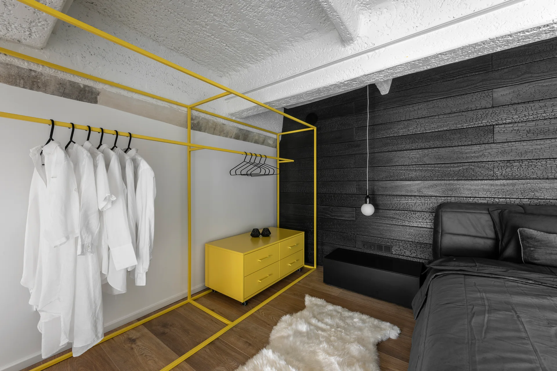 A modern bedroom features a bright yellow metal clothing rack and dresser set against a charred black wood accent wall and white concrete ceiling beams.