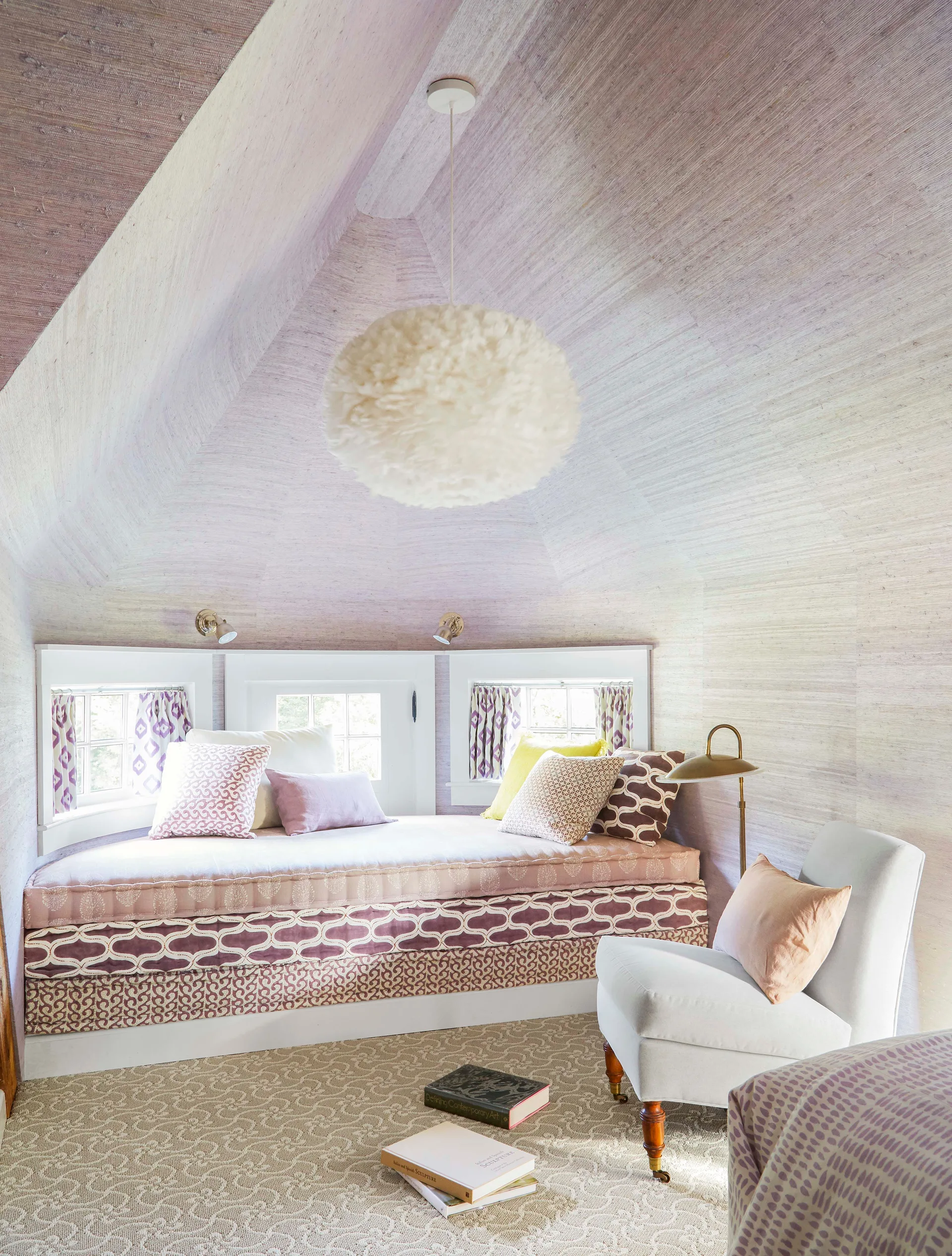 An attic bedroom nook features a built-in daybed with patterned pillows, lavender grasscloth walls, a fluffy pendant light, and a light grey armchair.