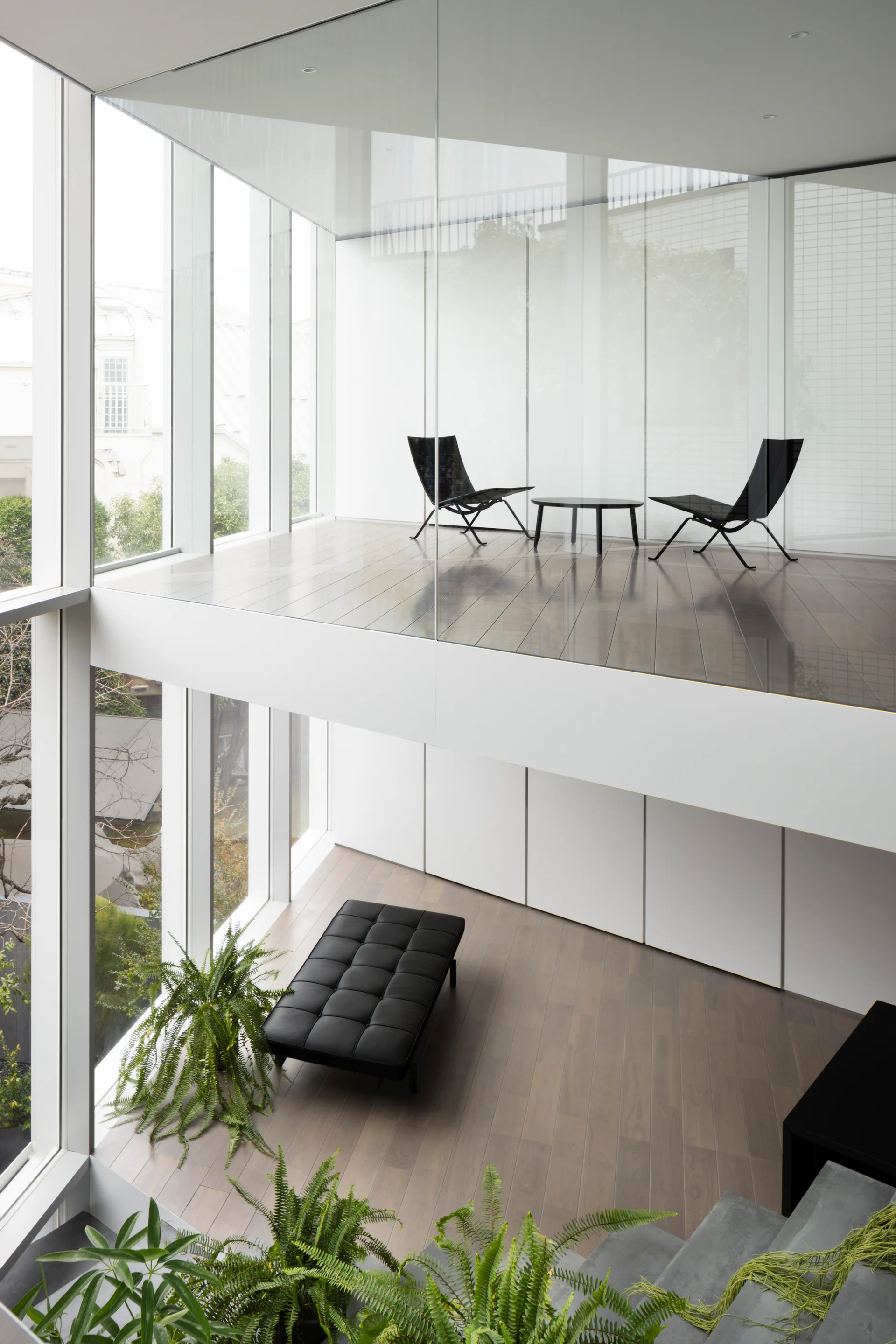 A double-height minimalist room features a glass-walled mezzanine with two black chairs, a lower level with a black leather bench, and green plants.
