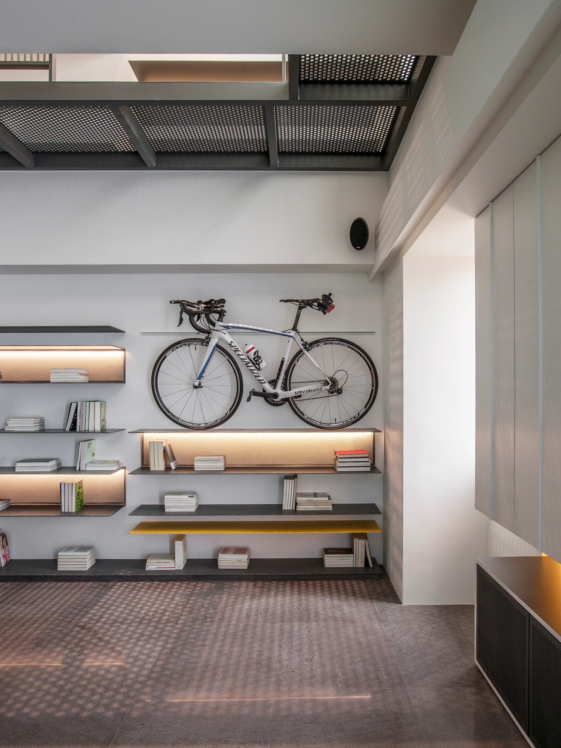A modern room featuring white walls with floating black shelves displaying books, a wall-mounted road bike, and a dark metal grid ceiling.