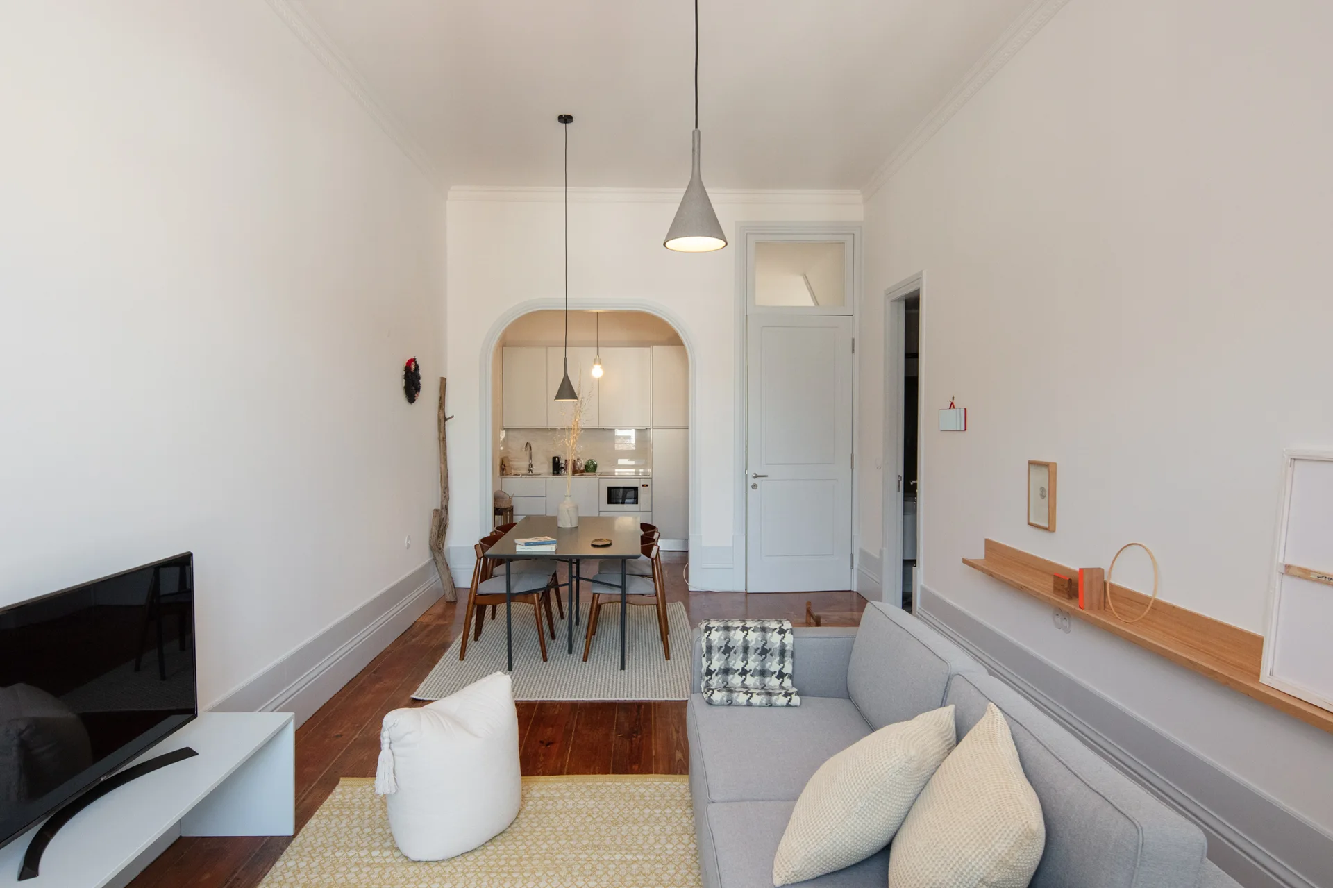 A minimalist living room features a gray sofa, a long wood shelf, and an arched doorway leading to a dining area.