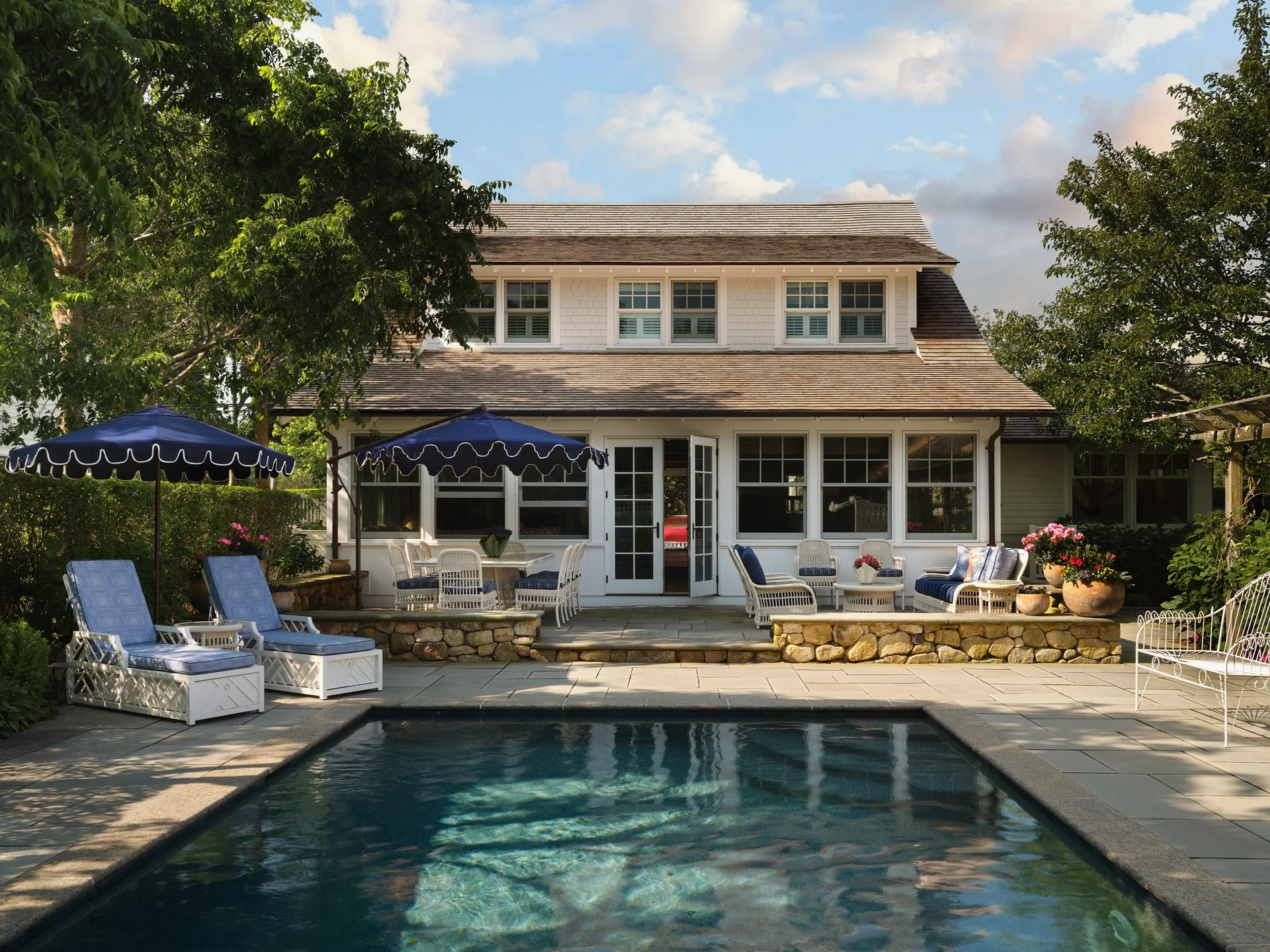A stone-paved patio with a rectangular swimming pool, blue and white lounge chairs, and a white shingled two-story house.
