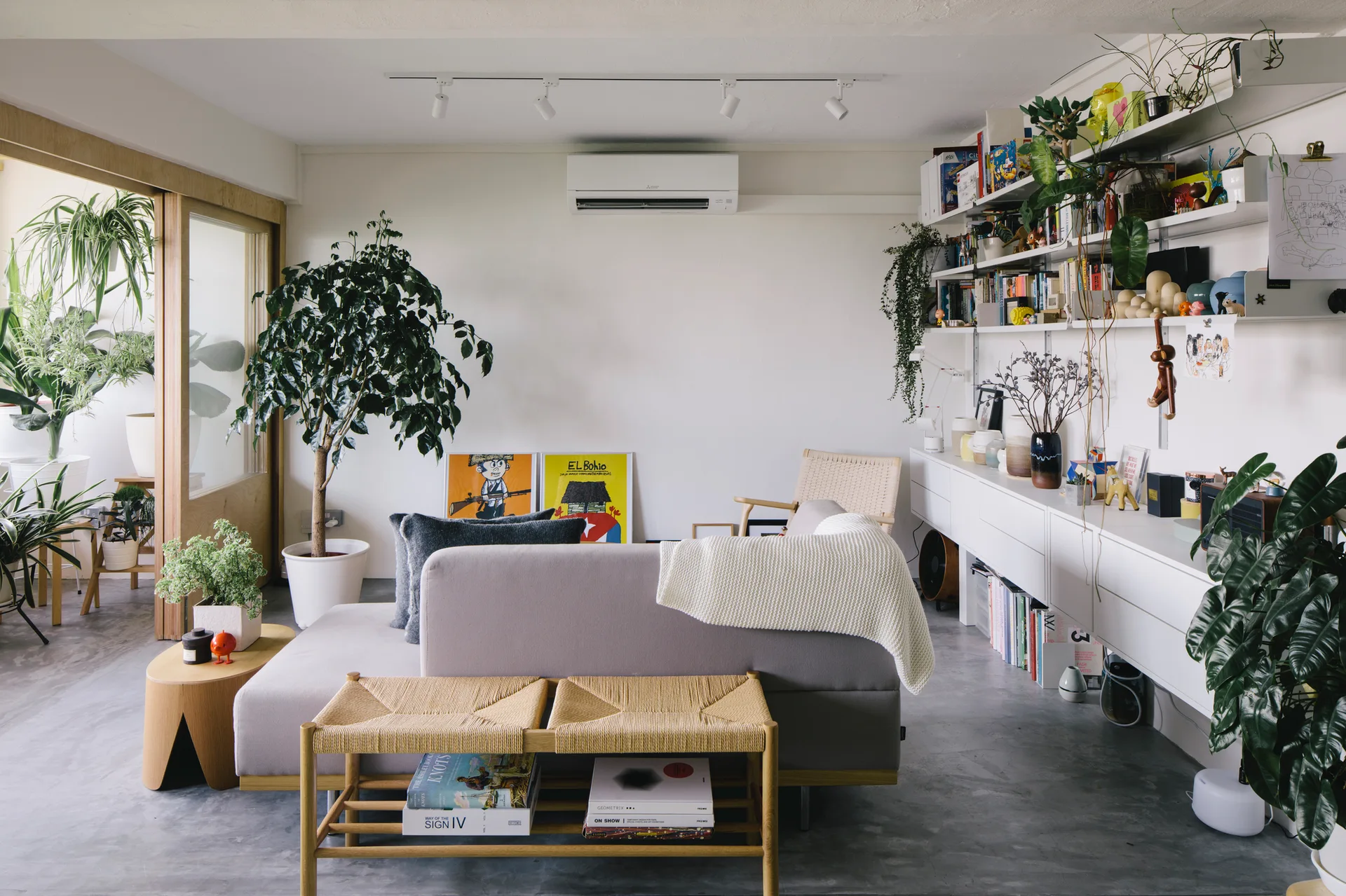 A bright living room features a grey sofa, a woven paper cord bench, white wall shelving filled with books, and potted plants on concrete floors.