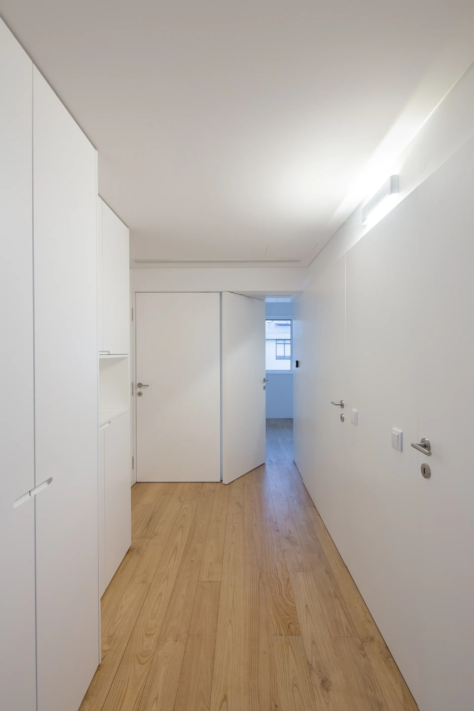 A minimalist hallway with white floor-to-ceiling cabinets, flush doors, linear ceiling lighting, and light wood floors.