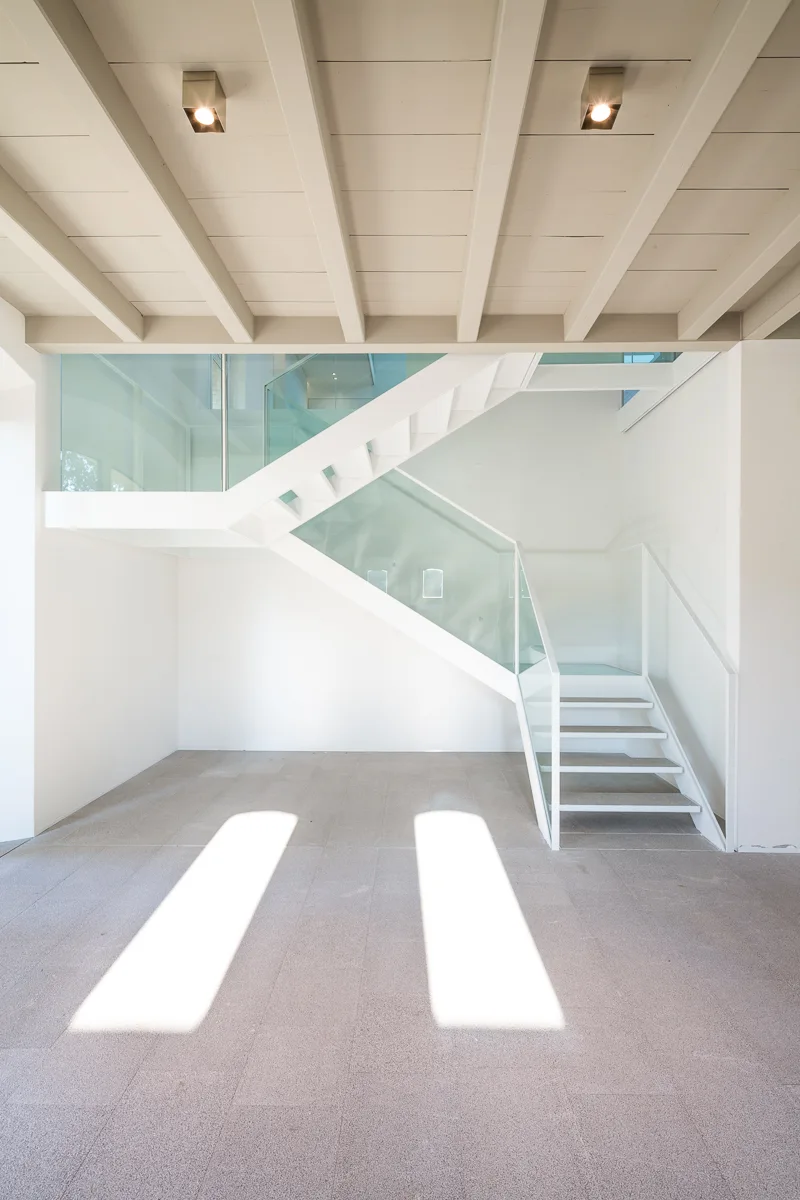 A modern white staircase with clear glass railings stands in an open area with exposed ceiling beams and light grey stone tile flooring.