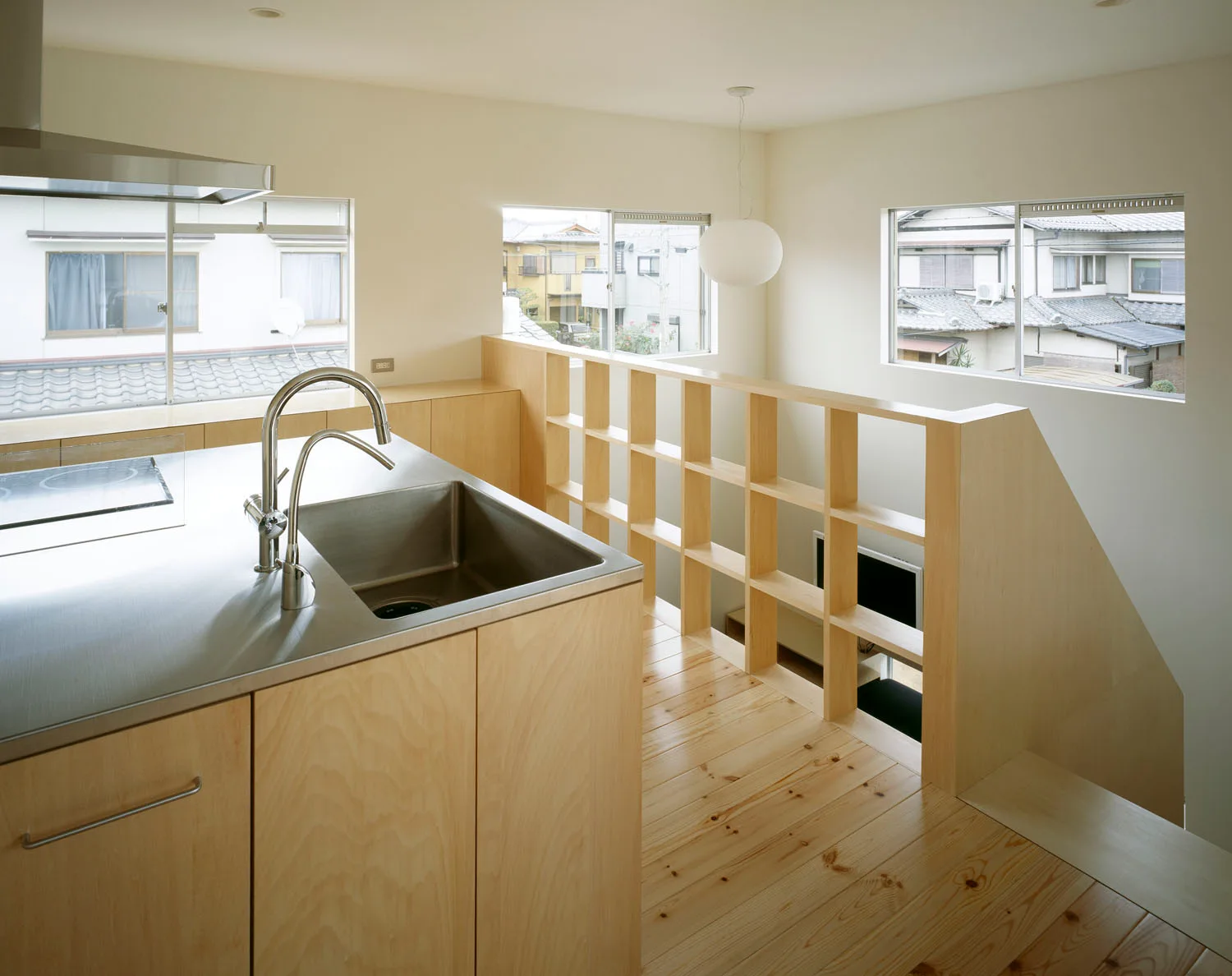 A minimalist kitchen features a stainless steel island with an integrated sink alongside light plywood cabinets and a wooden grid staircase railing.