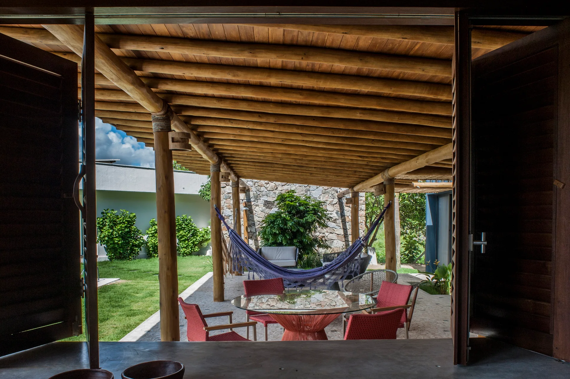 A covered patio with a rustic log roof features a circular glass dining table, red chairs, and a hammock in front of a stone wall.