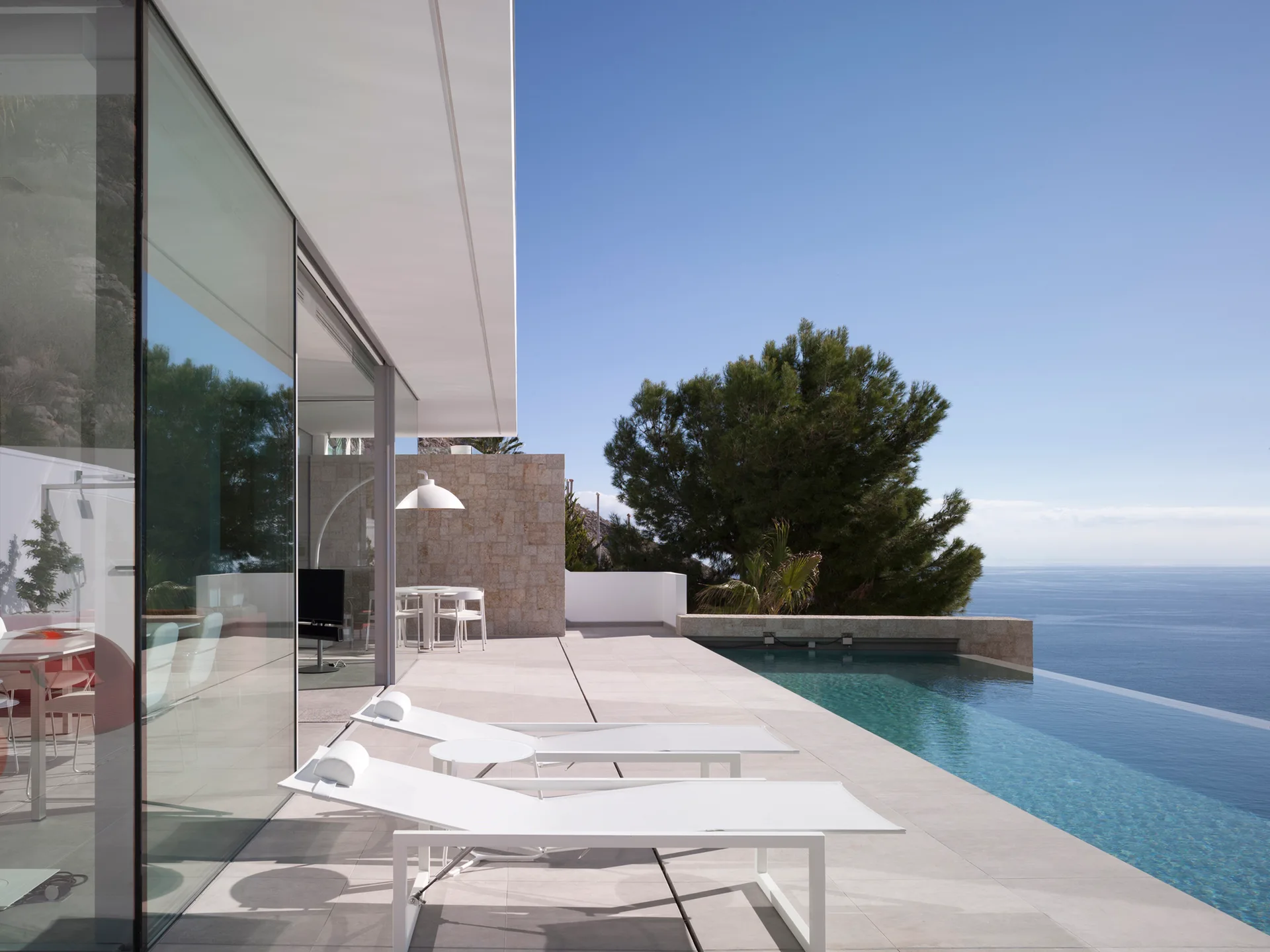 Two white sun loungers sit on a paved modern patio next to an infinity pool overlooking the ocean.