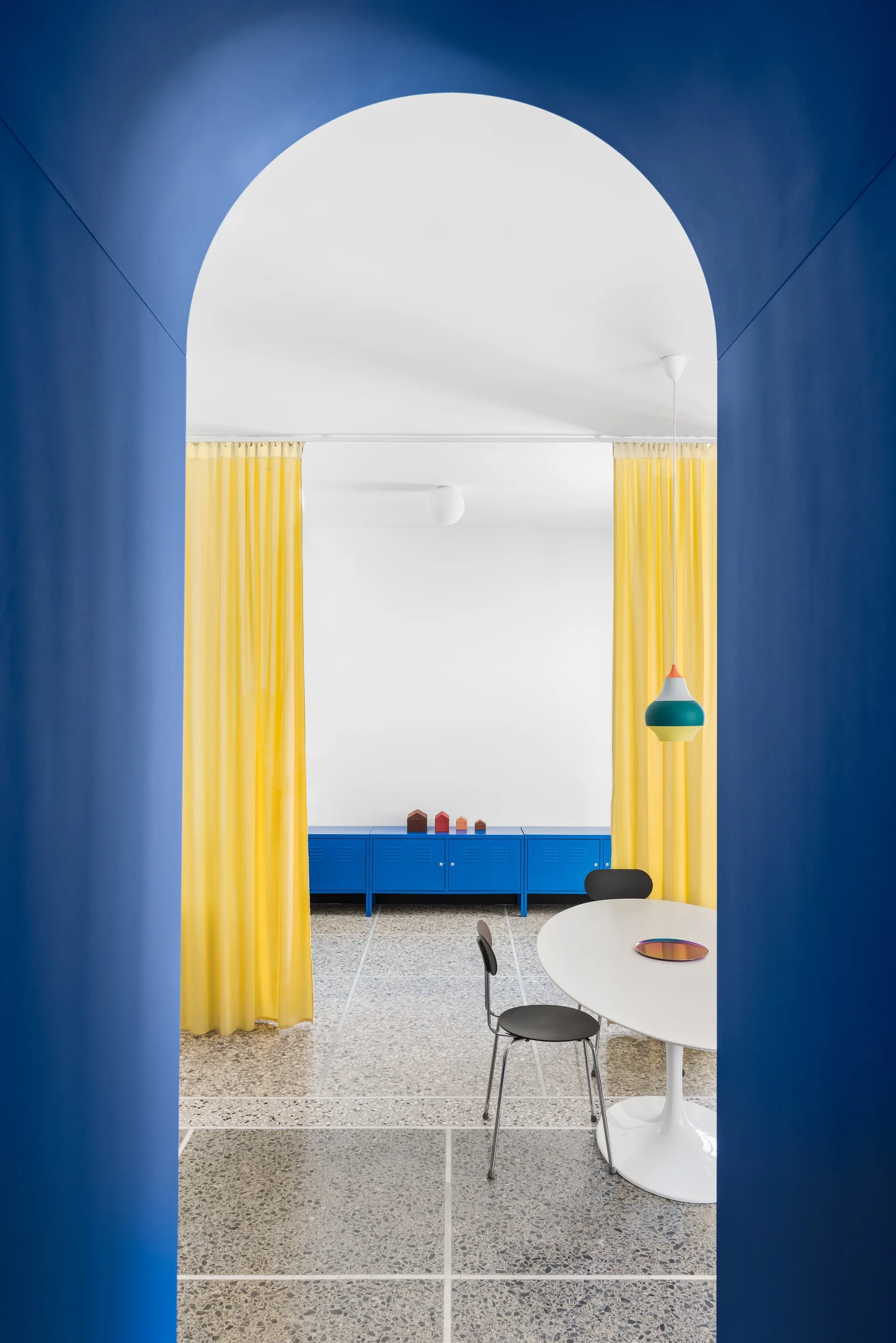 A dining room with a white pedestal table, yellow curtains, and a blue metal sideboard is viewed through a painted blue archway.