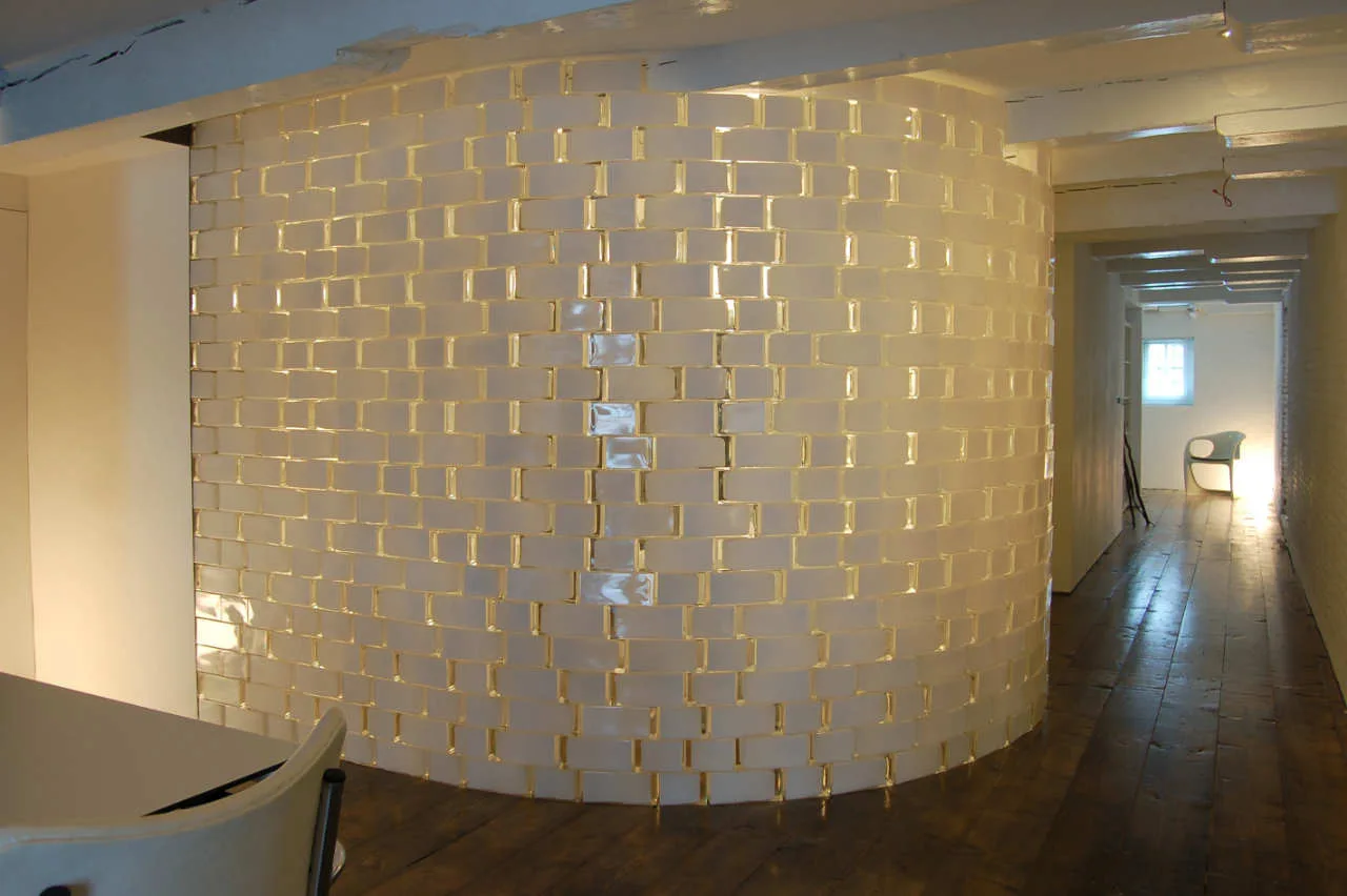 A curved partition wall built from glowing white translucent blocks divides a room with dark wood flooring and exposed ceiling beams.
