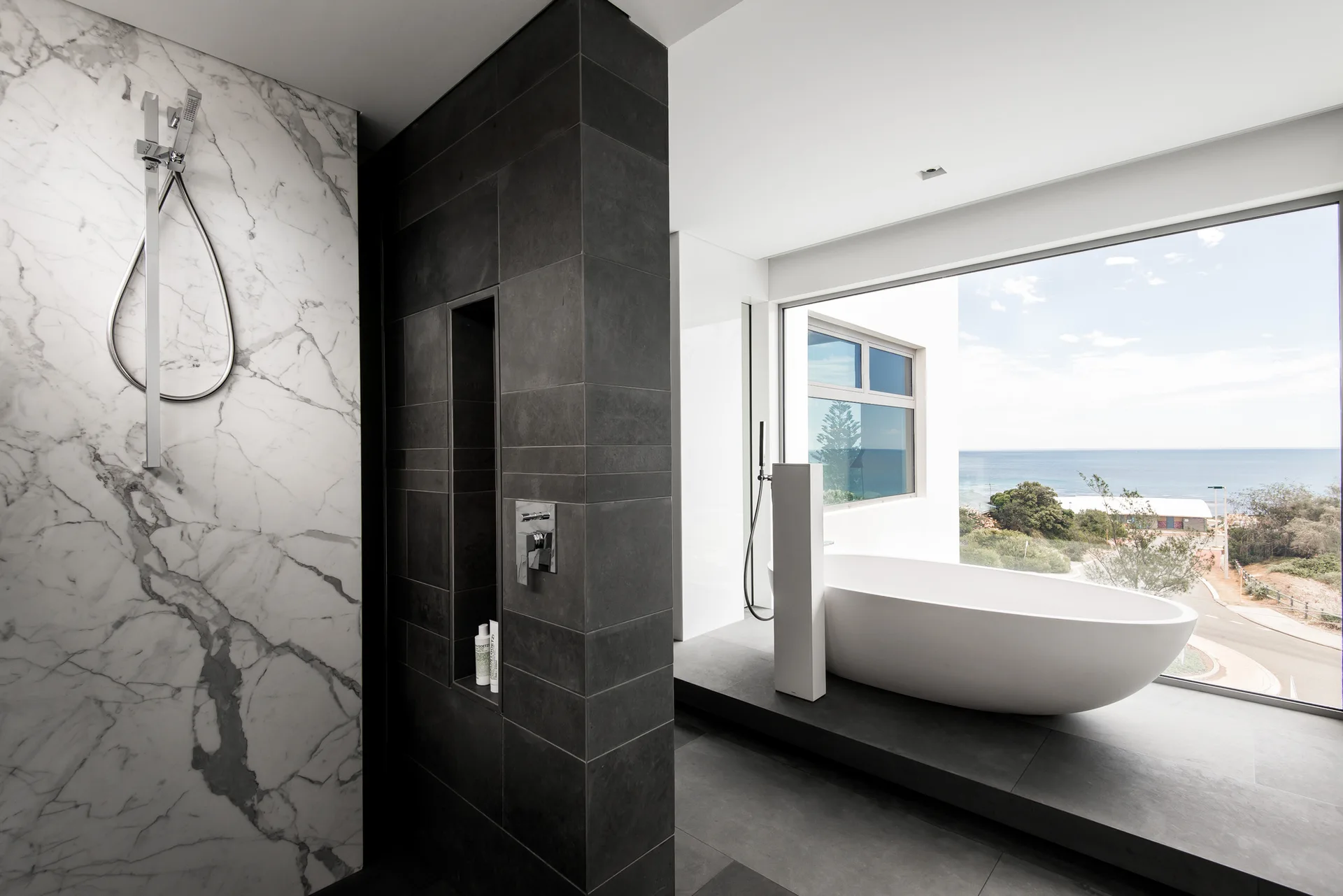 A modern bathroom featuring a freestanding white bathtub next to a window with ocean views and a walk-in shower with veined marble walls.