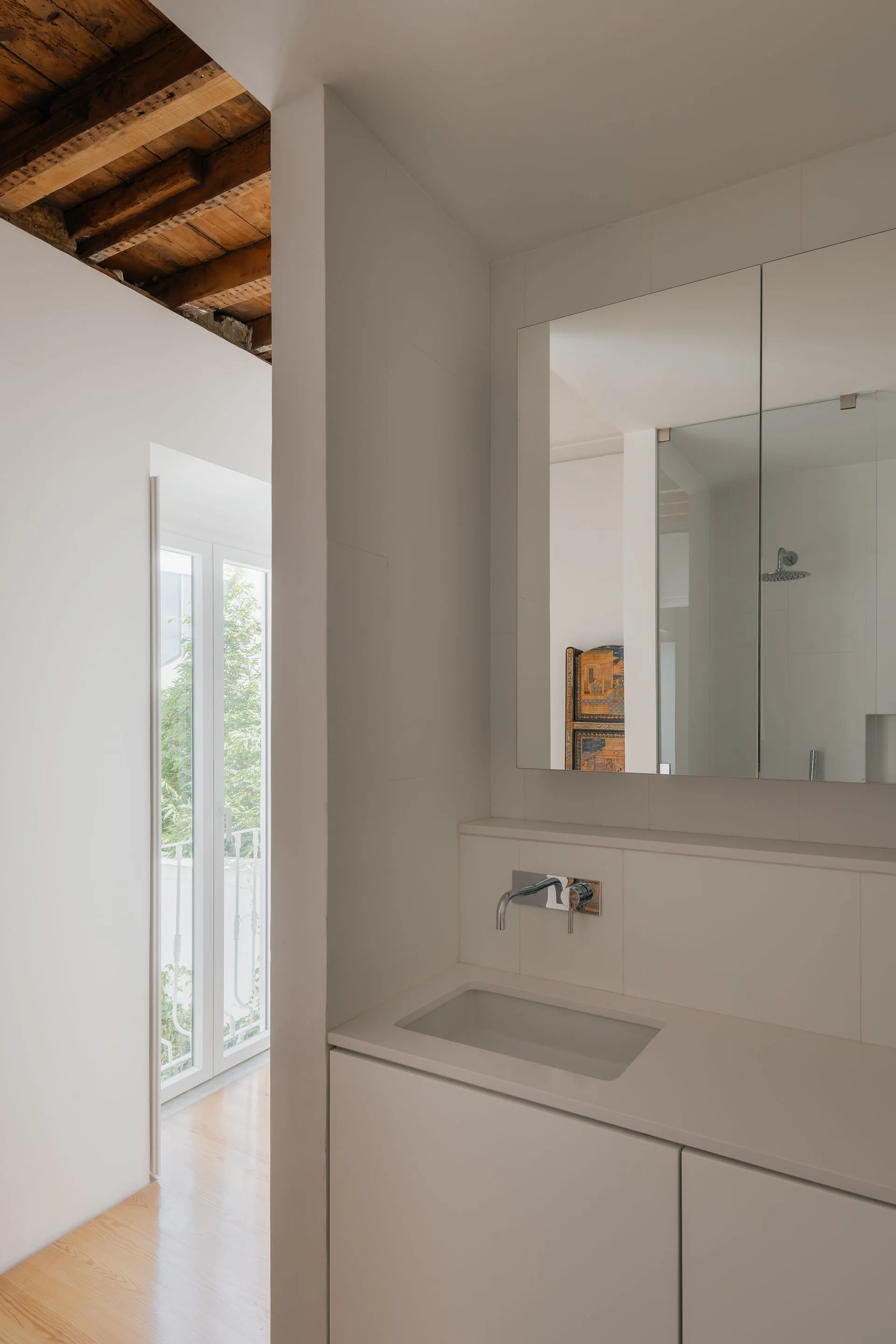 A minimalist bathroom features a white integrated sink vanity, a wall-mounted chrome faucet, a mirrored cabinet, and rustic wooden ceiling beams.