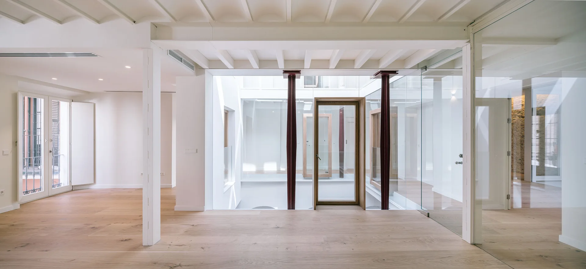 A minimalist interior corridor features light wood floors, an exposed beam ceiling, and a central glass-enclosed atrium supported by dark red steel columns.