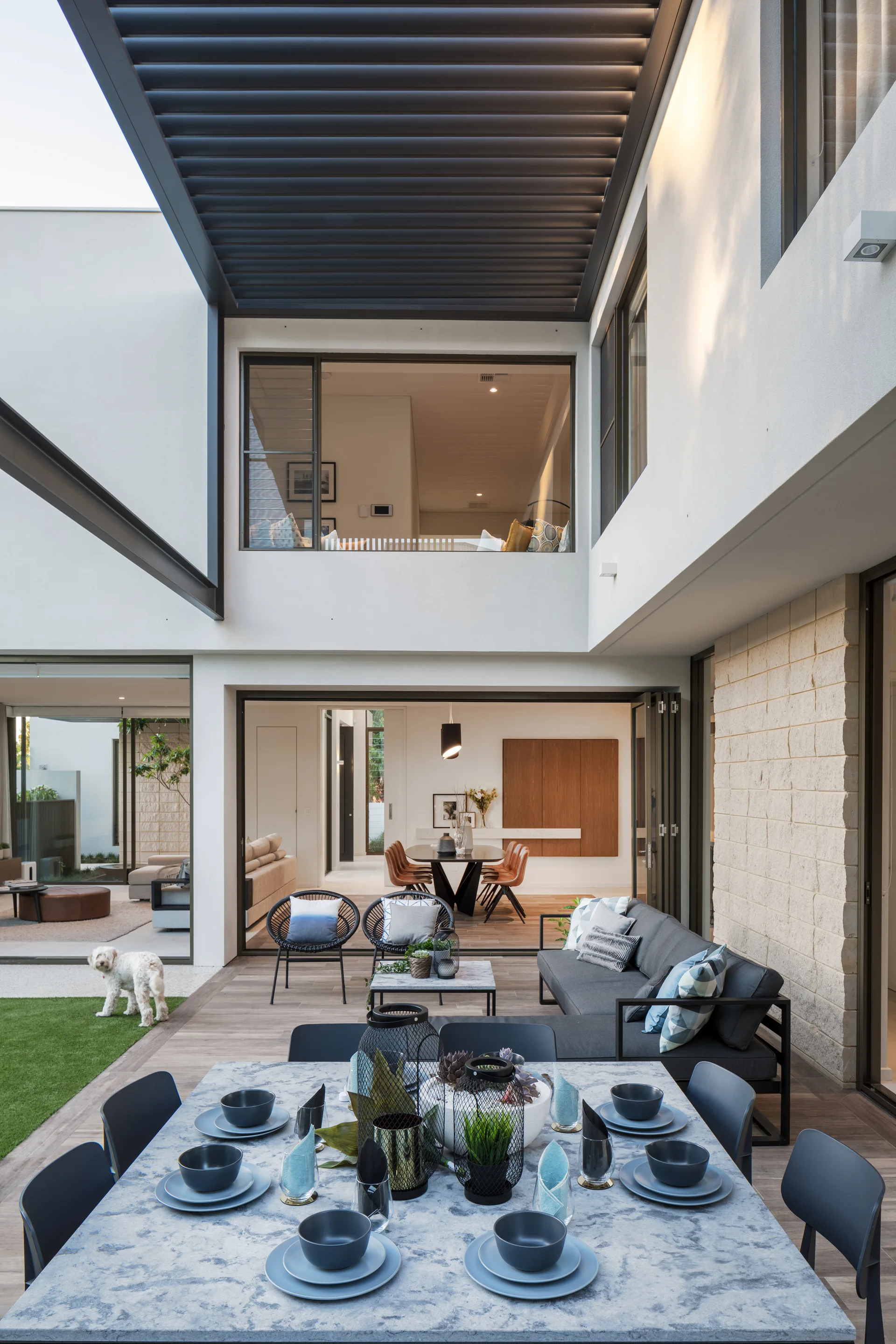 A modern outdoor patio features a black slatted pergola, a stone-top dining table with blue place settings, and a grey lounge sofa.