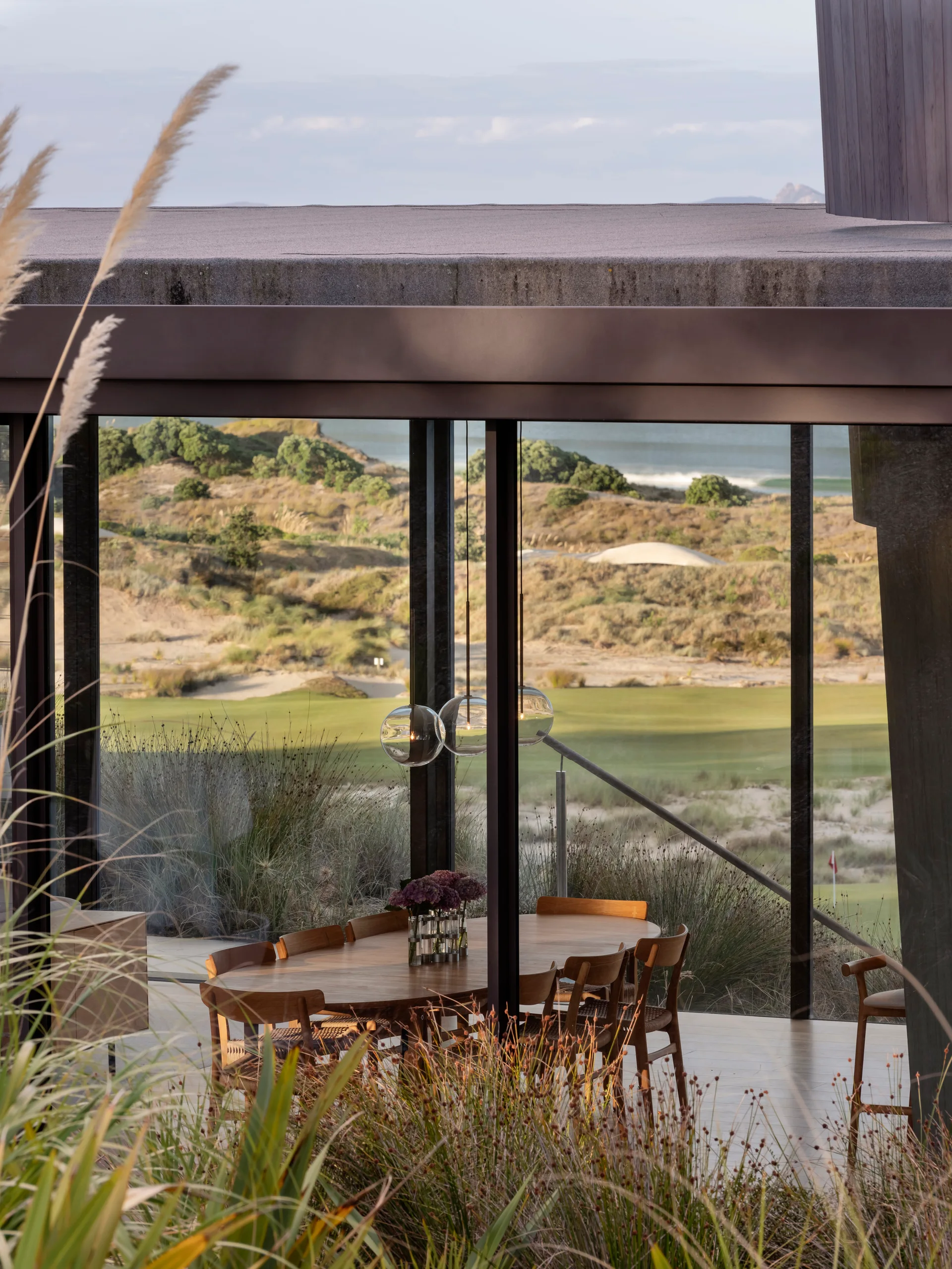 An oval wooden dining table surrounded by matching chairs sits before a large glass wall displaying a grassy dune landscape.