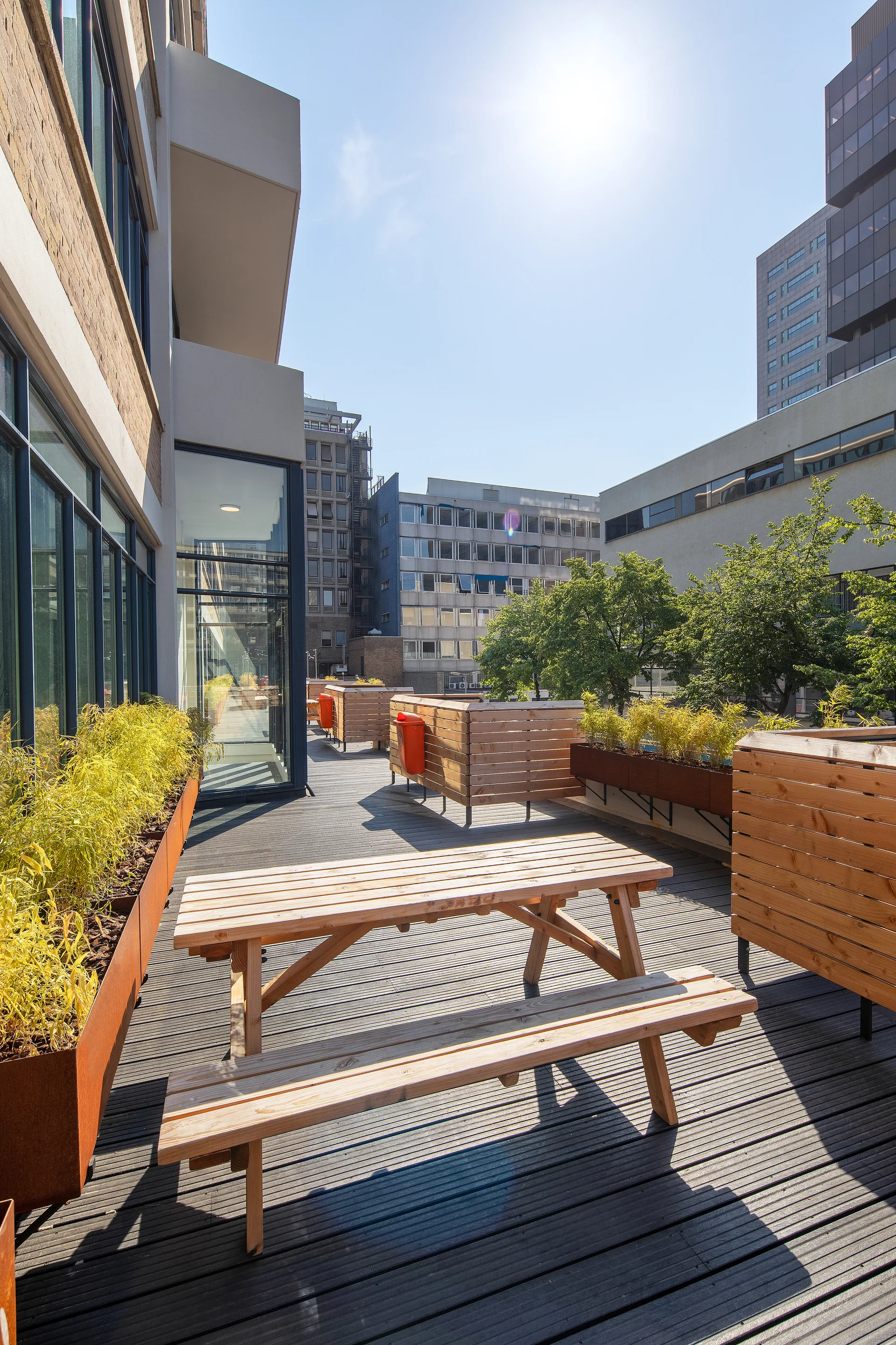 An urban rooftop terrace features a wooden picnic table on dark plank decking surrounded by slatted wood privacy screens and planters.