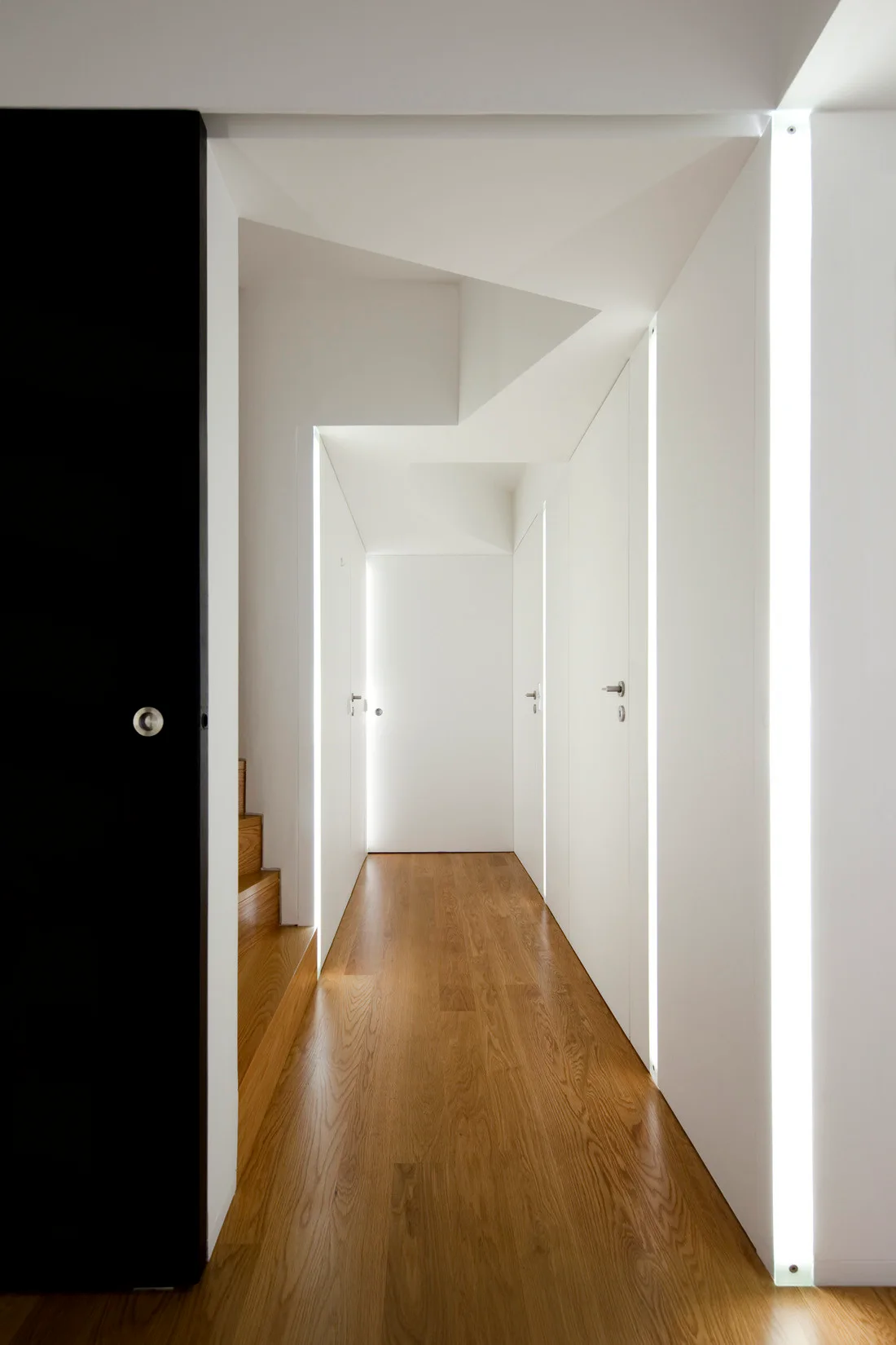 A minimalist hallway with white walls, natural wood floors, integrated vertical light strips beside flush doors, and a partial view of wooden stairs.