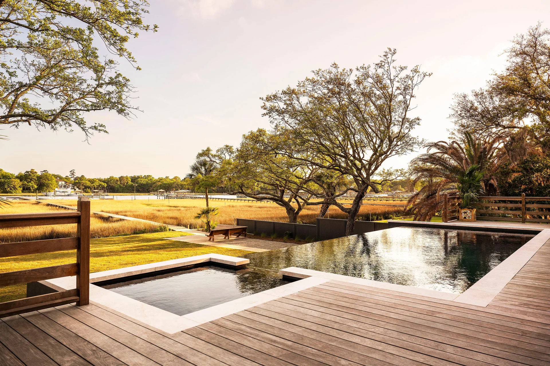 A rectangular infinity pool and integrated square spa set on a light brown wooden deck overlooking a grassy marshland with trees.