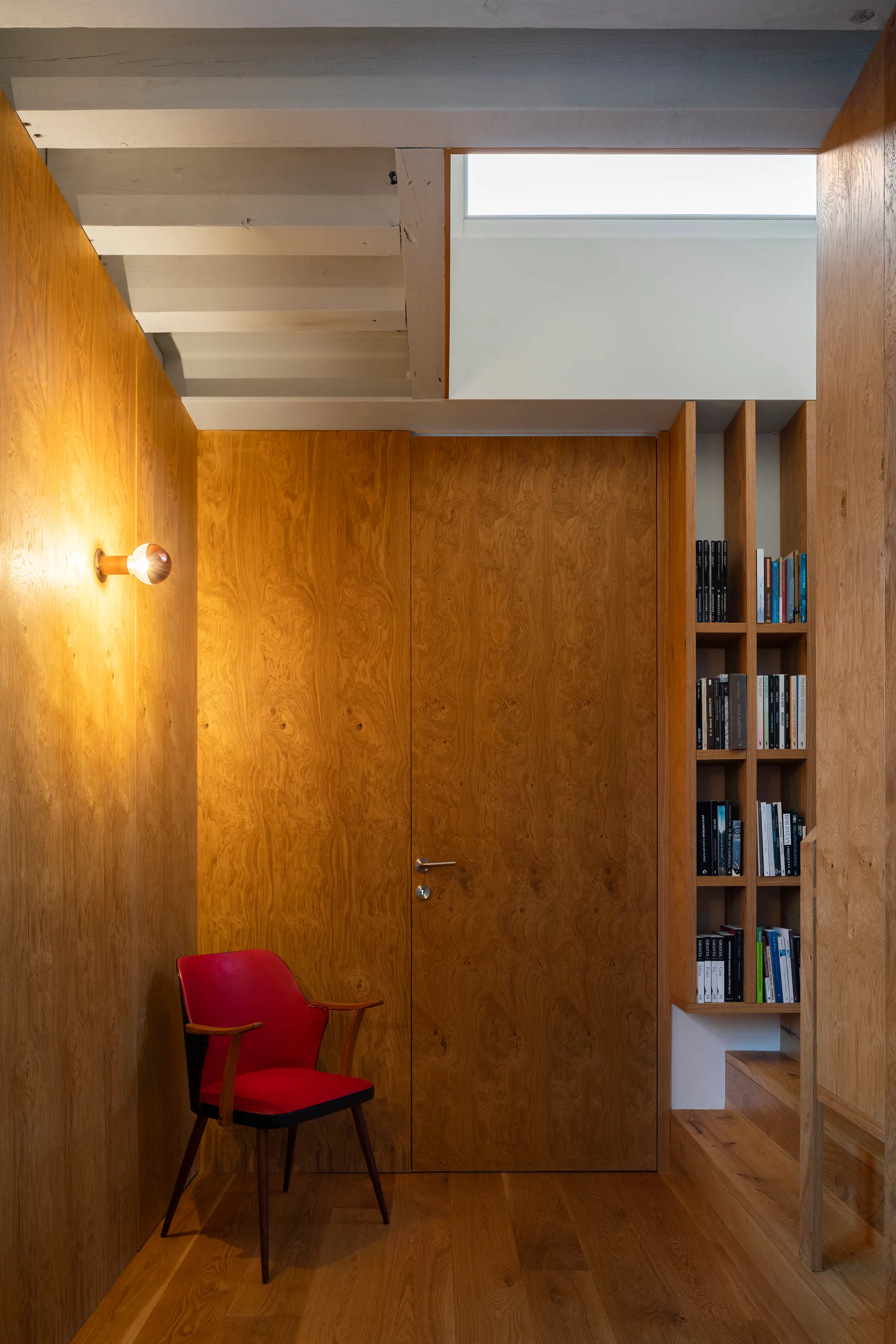 A wood-paneled hallway features a flush door, a single red accent chair, a round wall sconce, and a built-in wooden bookshelf next to a staircase.