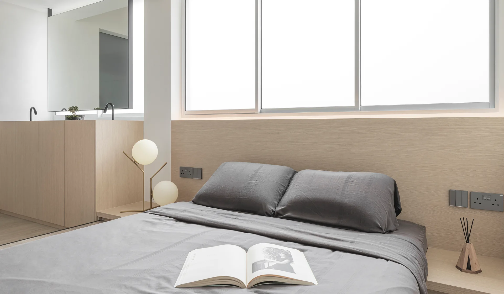A grey bed is set against a light wood integrated headboard wall with a matching bedside ledge, globe table lamp, and wide window.