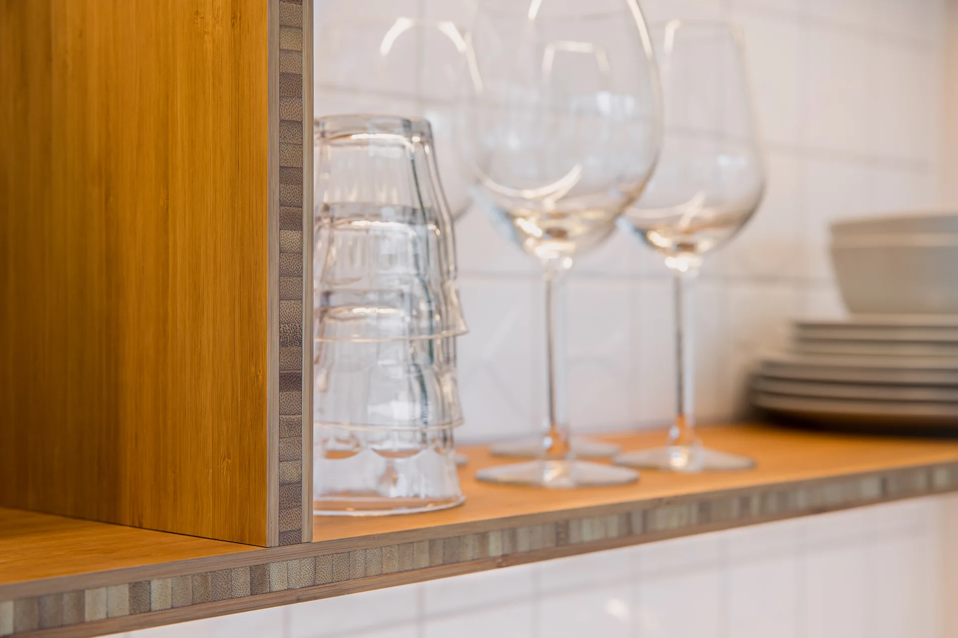 Stacked clear glasses and ceramic plates rest on a natural bamboo shelf in front of a white tiled kitchen backsplash.