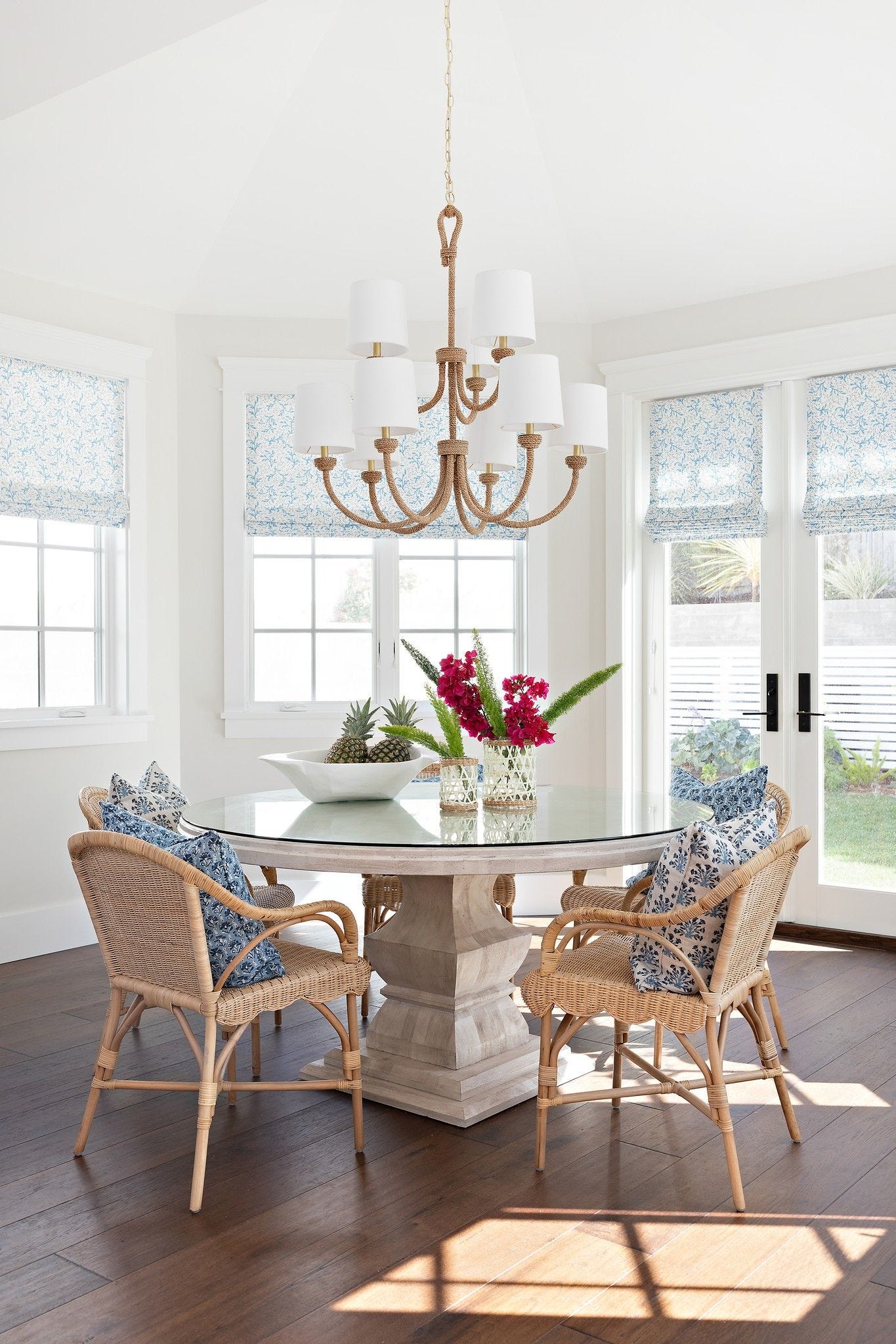 A coastal dining room features a round glass-top table, woven cane armchairs with blue-and-white patterned cushions, and a rope-wrapped chandelier.