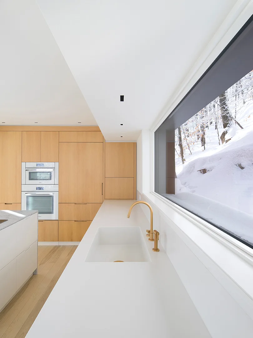 Modern kitchen with light wood flat-panel cabinets, a white countertop, a brass faucet, and a large window looking out onto snowy woods.